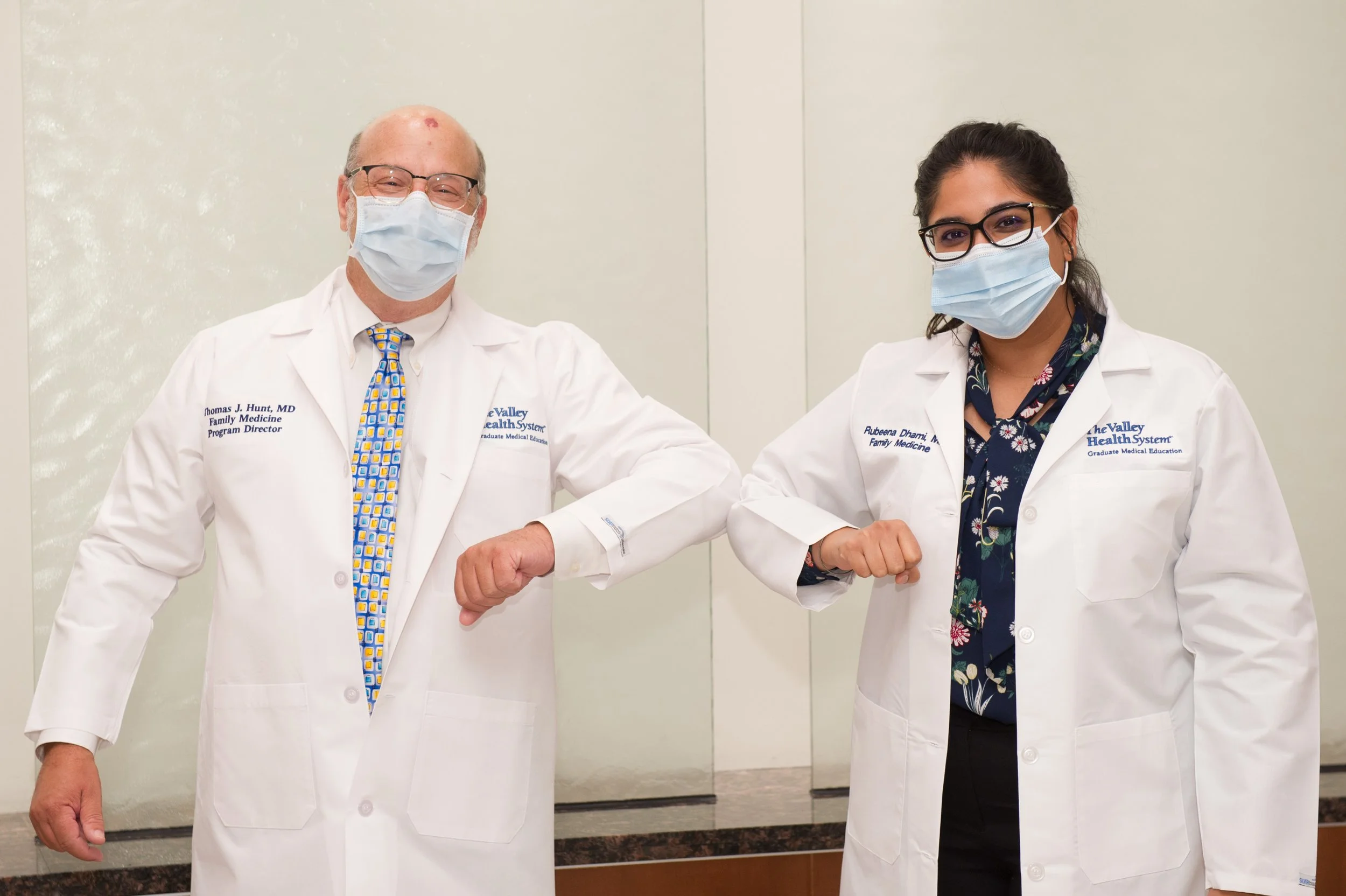 Dr. Dhami and her PD are wearing white coats and face masks, standing indoors against a plain wall, greeting each other with an elbow bump. The man has glasses and a colorful tie.  Dr. Dhami has glasses and a floral-patterned shirt under her lab coat