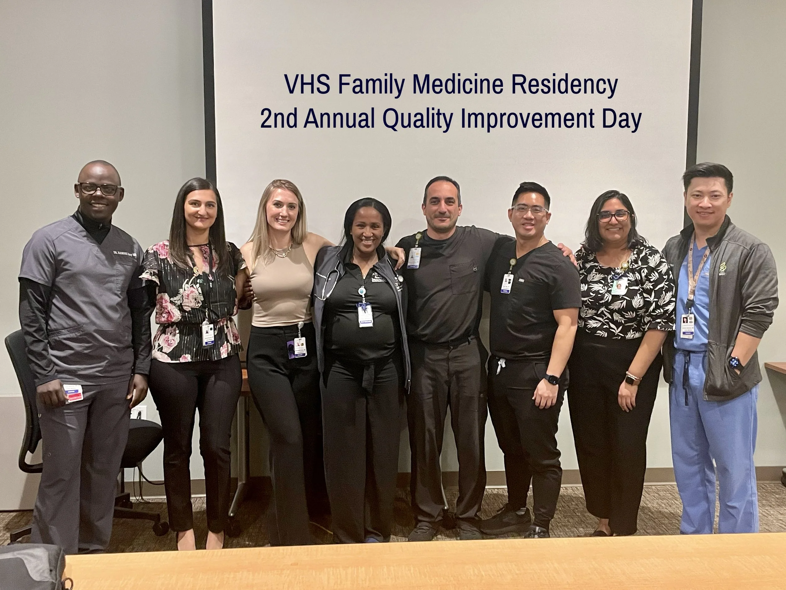 Dr. Dhami and her residency cohort standing together in front of a screen that reads 'VHS Family Medicine Residency 2nd Annual Quality Improvement Day'.