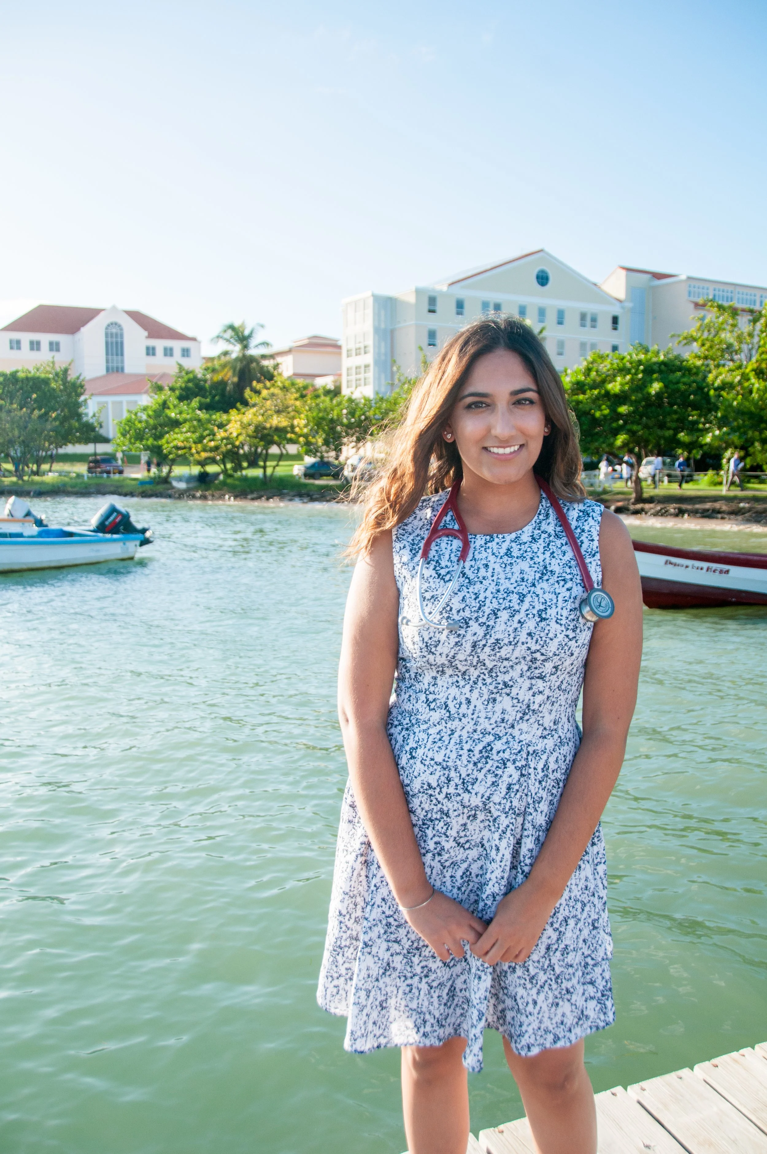 Dr. Rubeena Dhami, MD standing on a dock by the water, smiling, wearing a blue and white patterned dress, with a stethoscope around her neck, with buildings and trees of the St. George's University School of Medicine in the background.