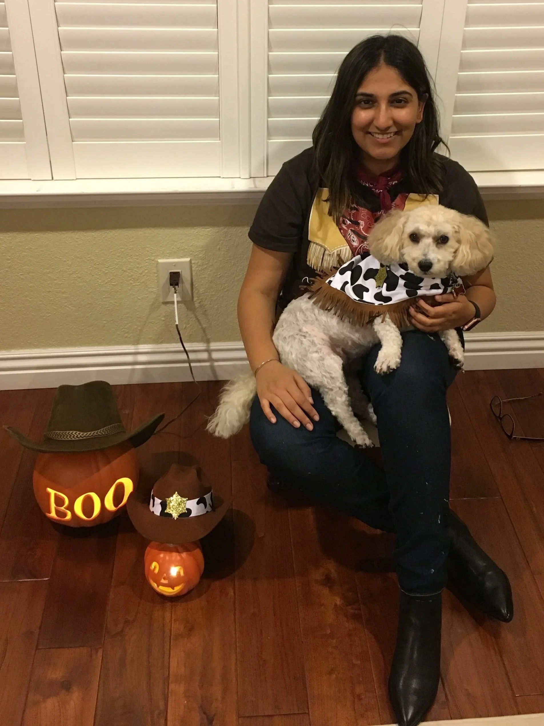 A woman with dark hair smiling while holding a small white dog in her lap. The woman is wearing a black shirt and a red bandana. The dog is dressed in a cow print bandana. In the background, there are white window shutters. On the wood floor, there are two carved pumpkin decorations, one with a jack-o'-lantern face and the other painted with the word "BOO"