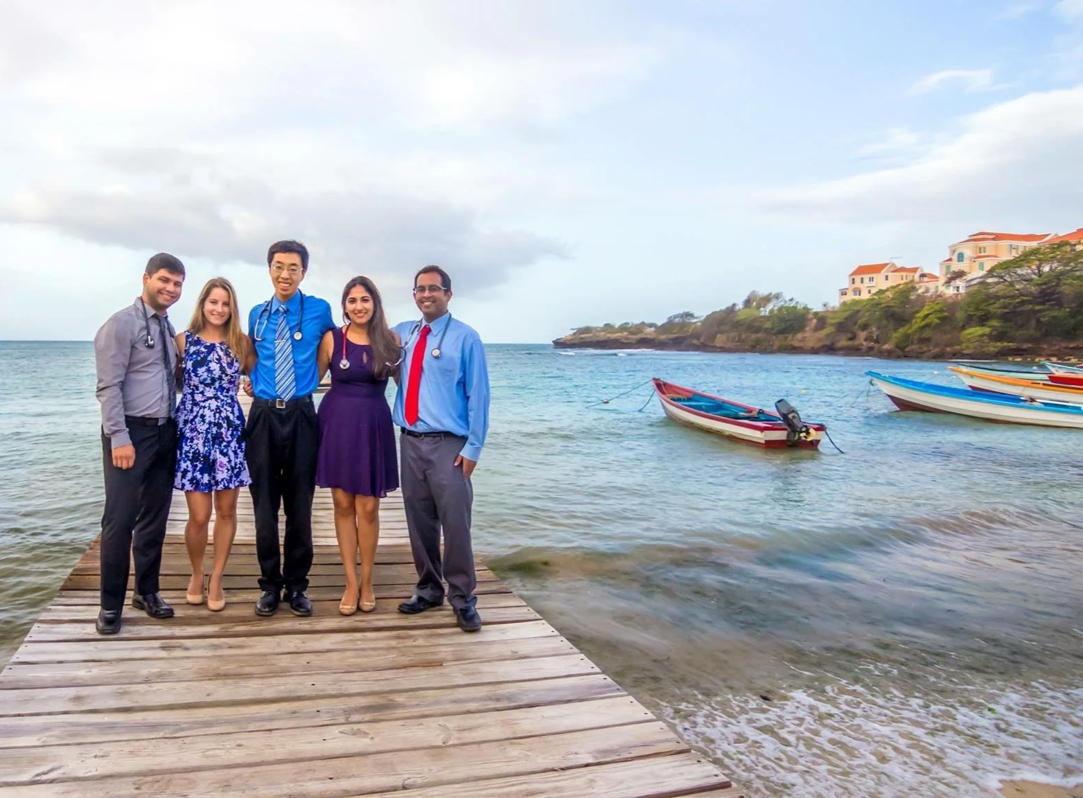 Dr. Dhami in a group of 5 medical students in business casual attire, standing on a wooden dock by the ocean with boats and houses on a hill in the background.