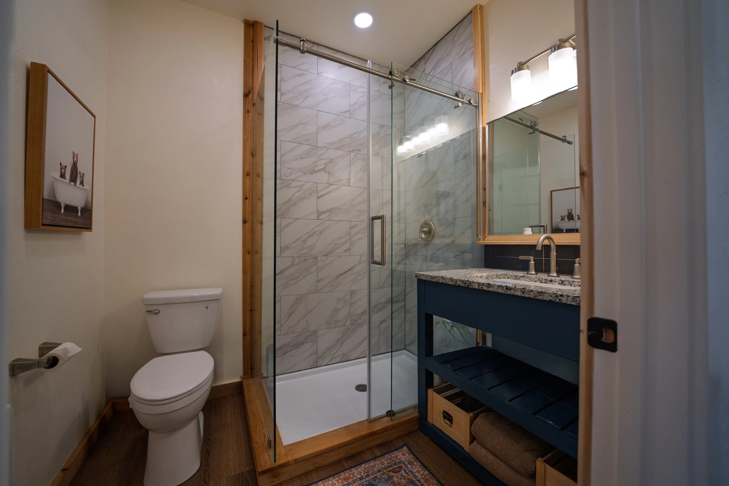 Bathroom with a toilet, a glass shower enclosure with marble tiles, a blue vanity with a granite countertop, a large mirror, and wall-mounted lights.