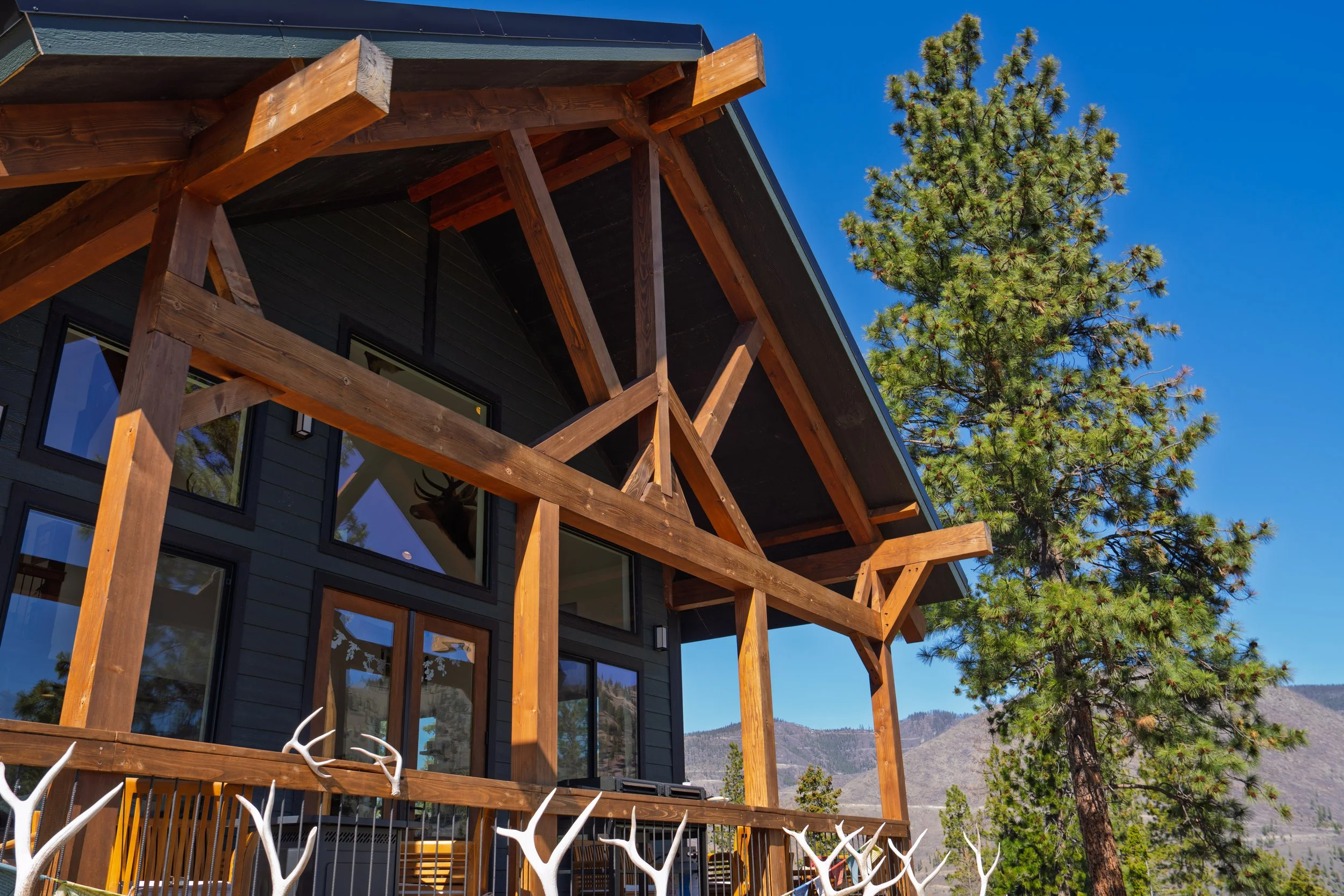 A modern house with a wooden balcony structure, large windows, and a dark exterior, surrounded by trees and mountains under a clear blue sky.