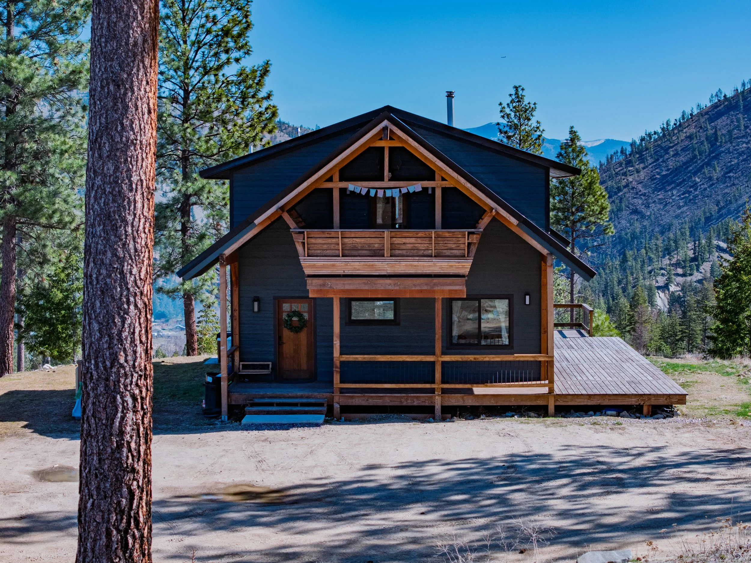 A dark blue wooden cabin with a sloped roof is situated in a forested mountain area, featuring a front porch, balcony, and decorative hanging flags.