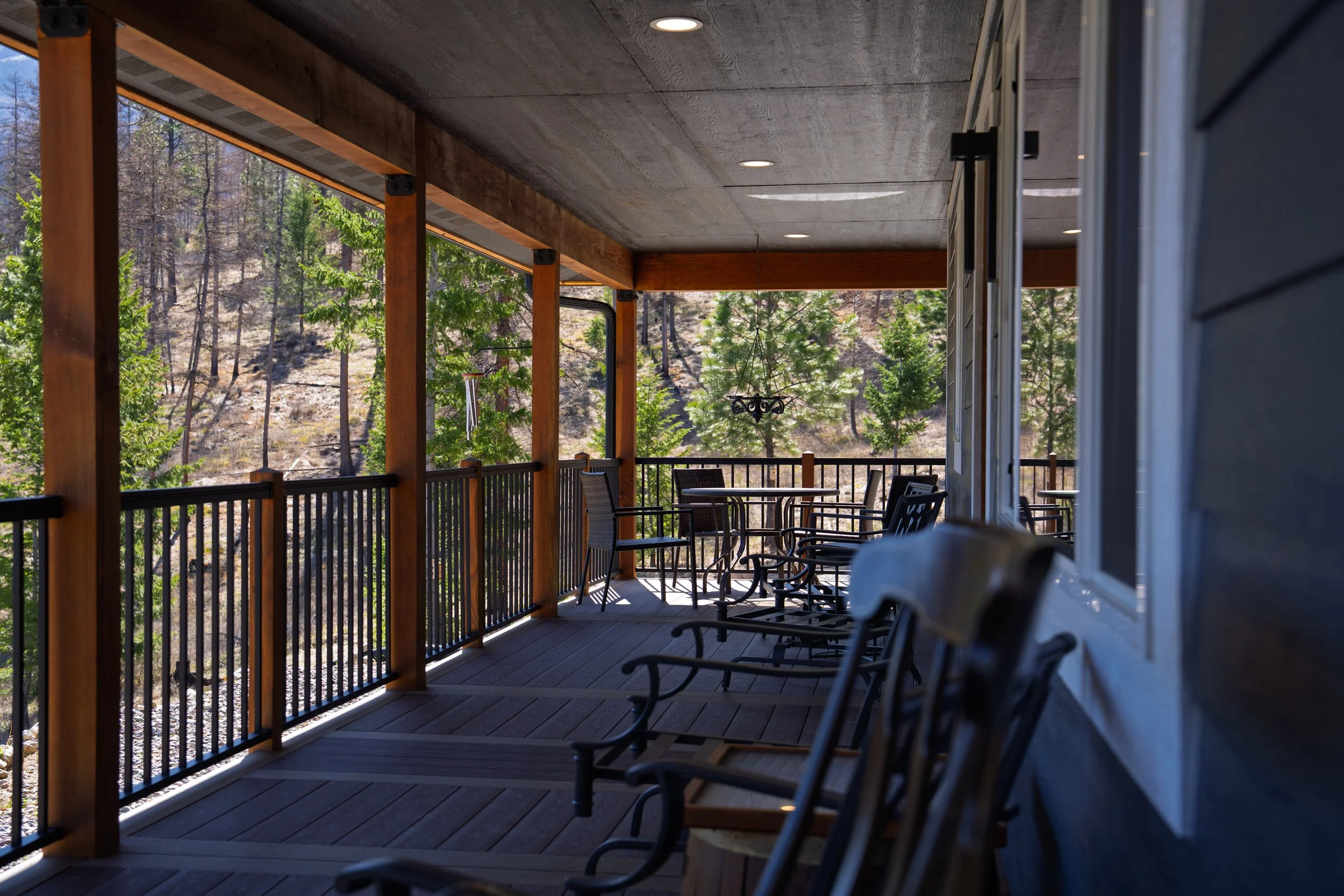 Covered outdoor balcony with wooden railing and black metal chairs and tables, overlooking a forested hillside with trees and rocks.