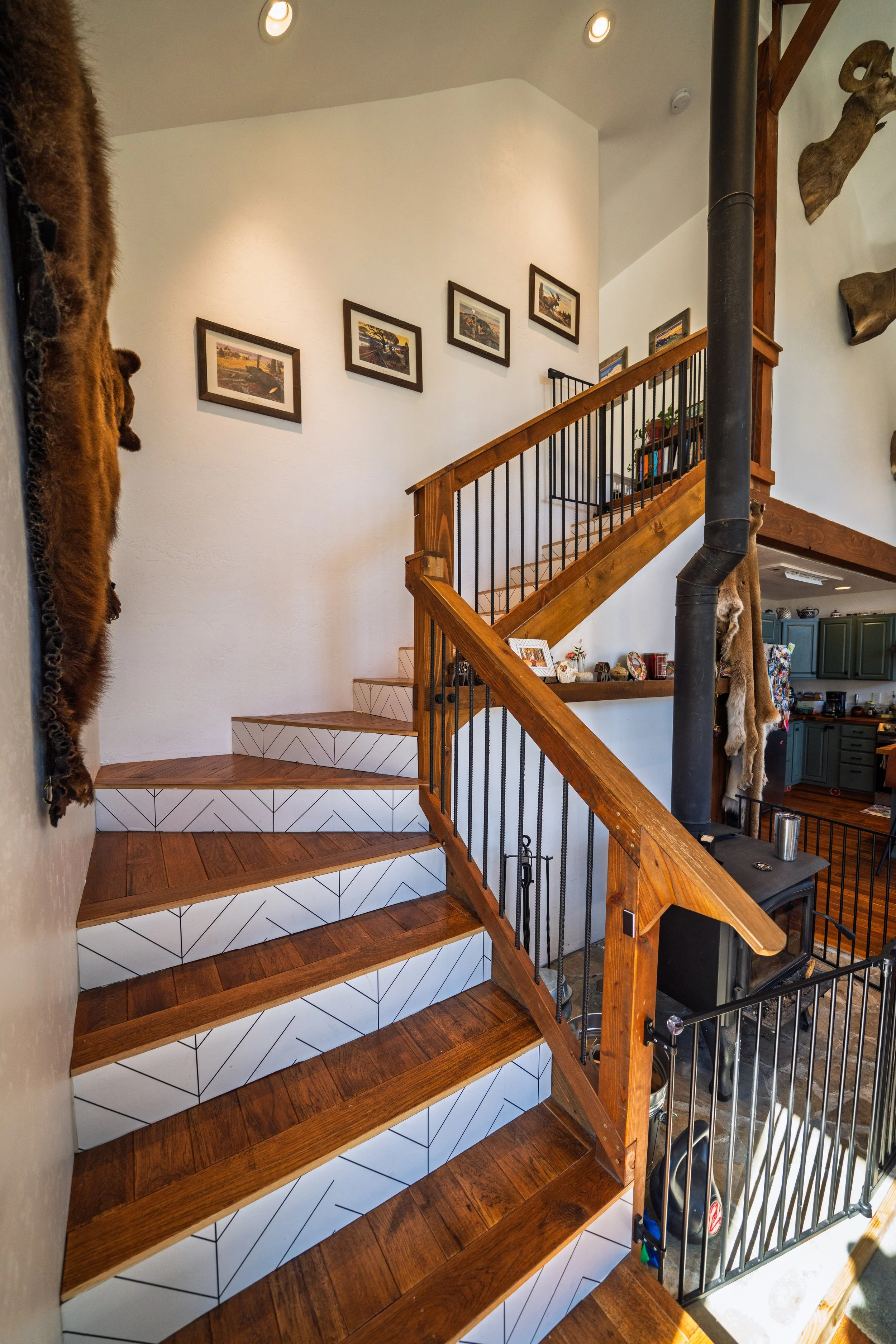 Wooden staircase with black metal railing, leading upstairs in a home with white walls decorated with landscape paintings and animal pelts on the wall.