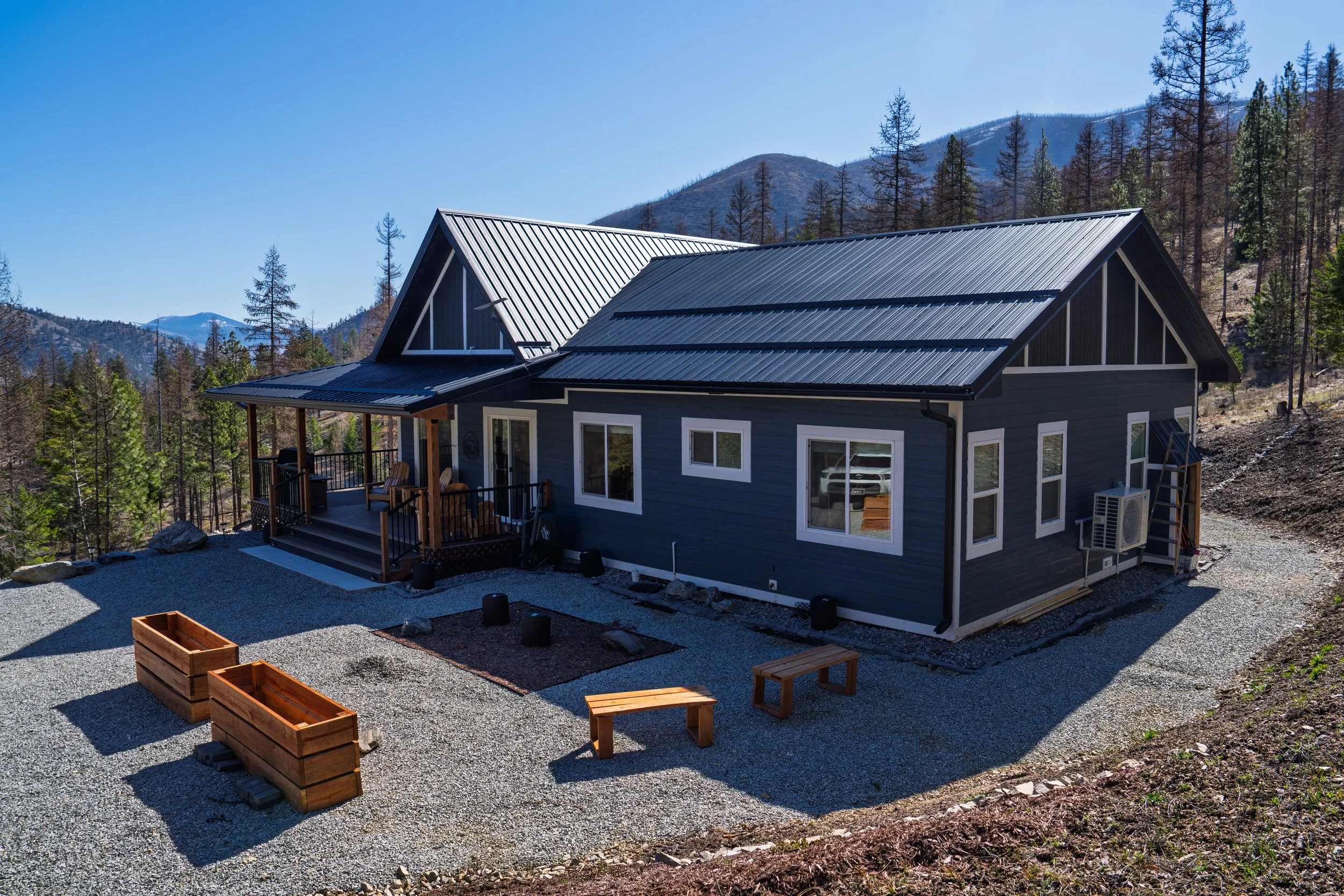 A modern black house with a metal roof, situated in a mountainous area with pine trees. It has a wooden porch with outdoor seating and a gravel yard with wooden planters and benches.
