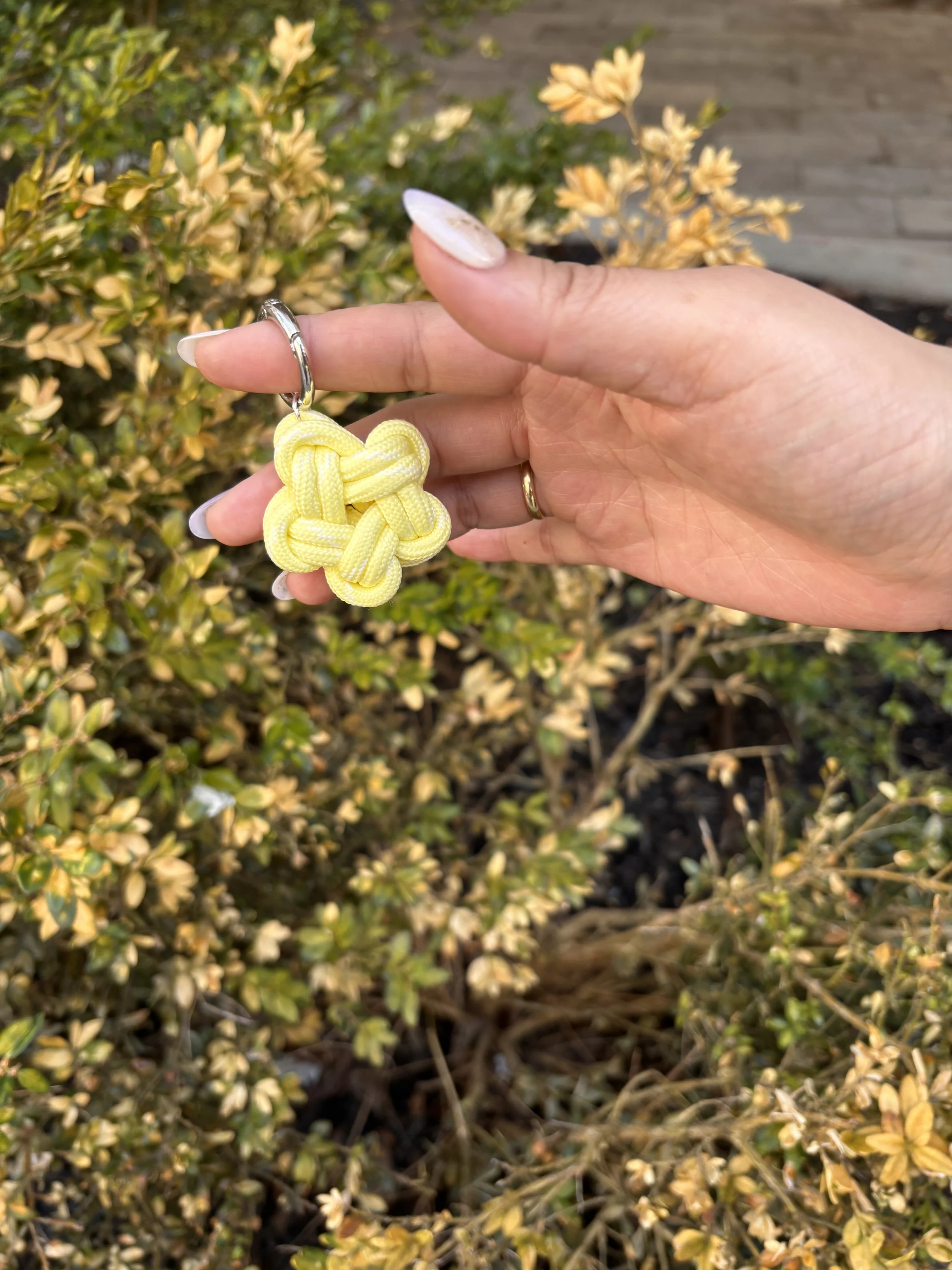 A hand holding a yellow paracord keychain in the shape of a knot, with a garden bush in the background.
