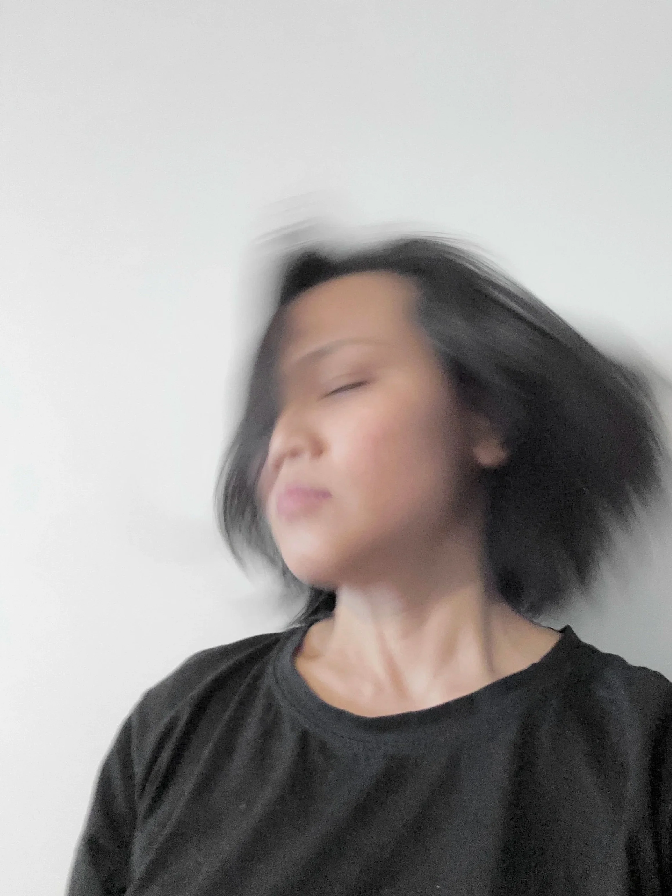 A woman with shoulder-length black hair wearing a black T-shirt, her head tilted slightly, with motion blur on her hair and face, against a plain white background.