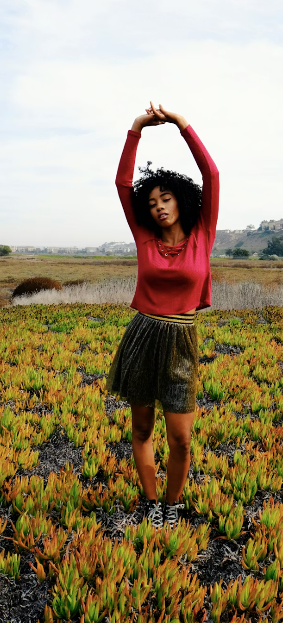 Young woman standing in a field of succulents with her arms raised above her head, wearing a red long sleeve shirt, a black pleated skirt, and sneakers, with hills and cloudy sky in the background.