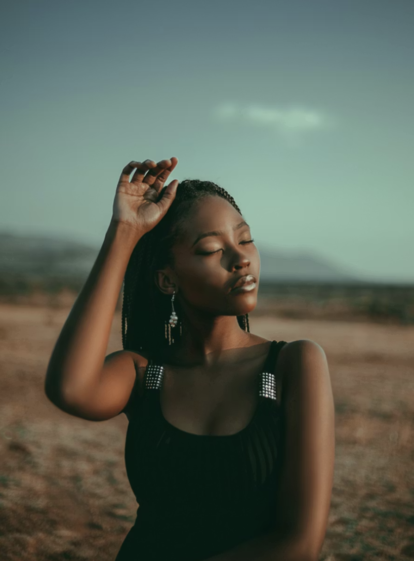 A woman with braided hair and earrings stands outdoors with her eyes closed, her right hand raised and touching her head, against a desert landscape with a clear sky.