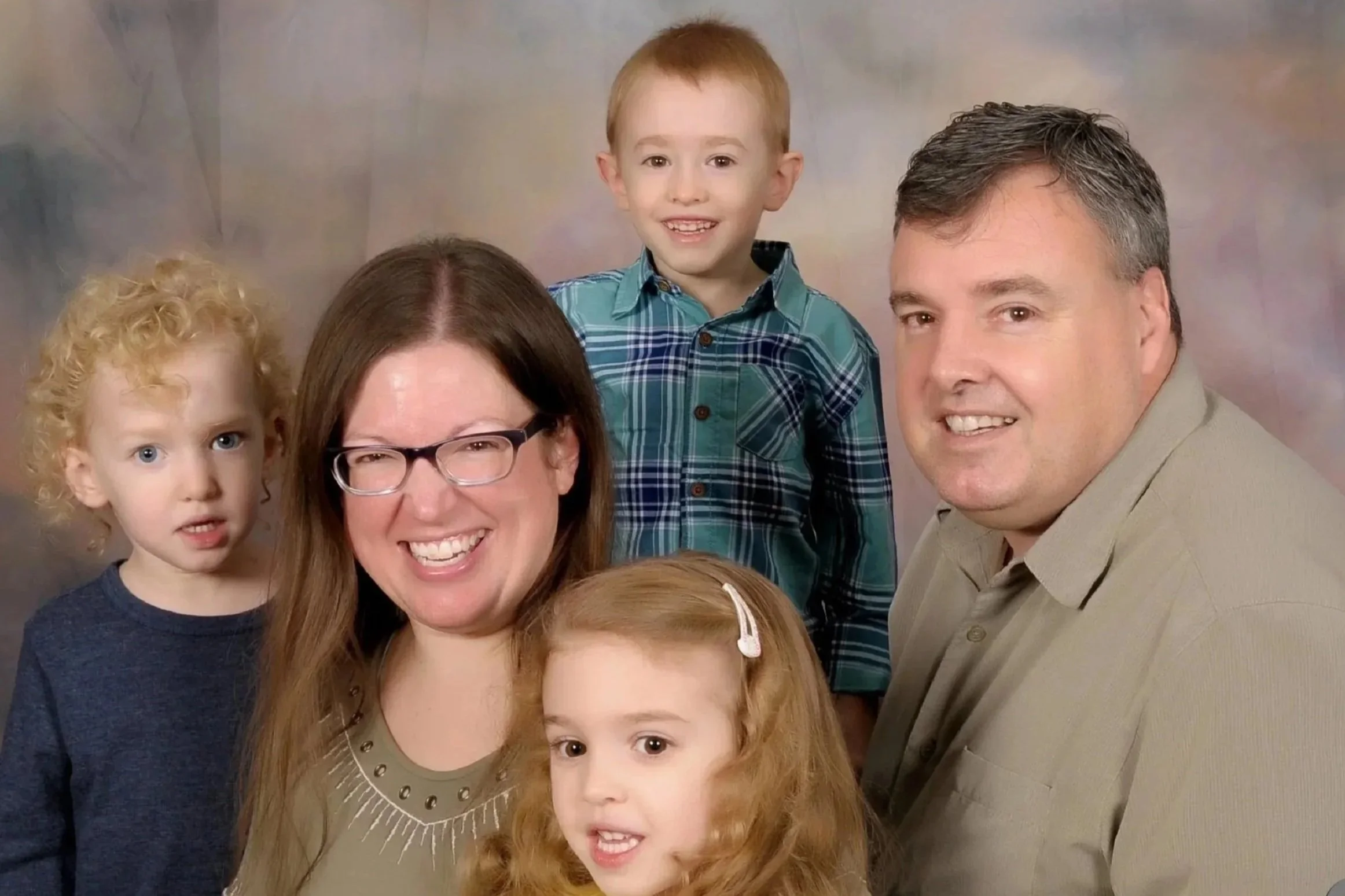 A family portrait featuring a woman with glasses, a man, and three children: a girl with red hair, a boy with short red hair, and a girl with curly blonde hair, all smiling and posing together.