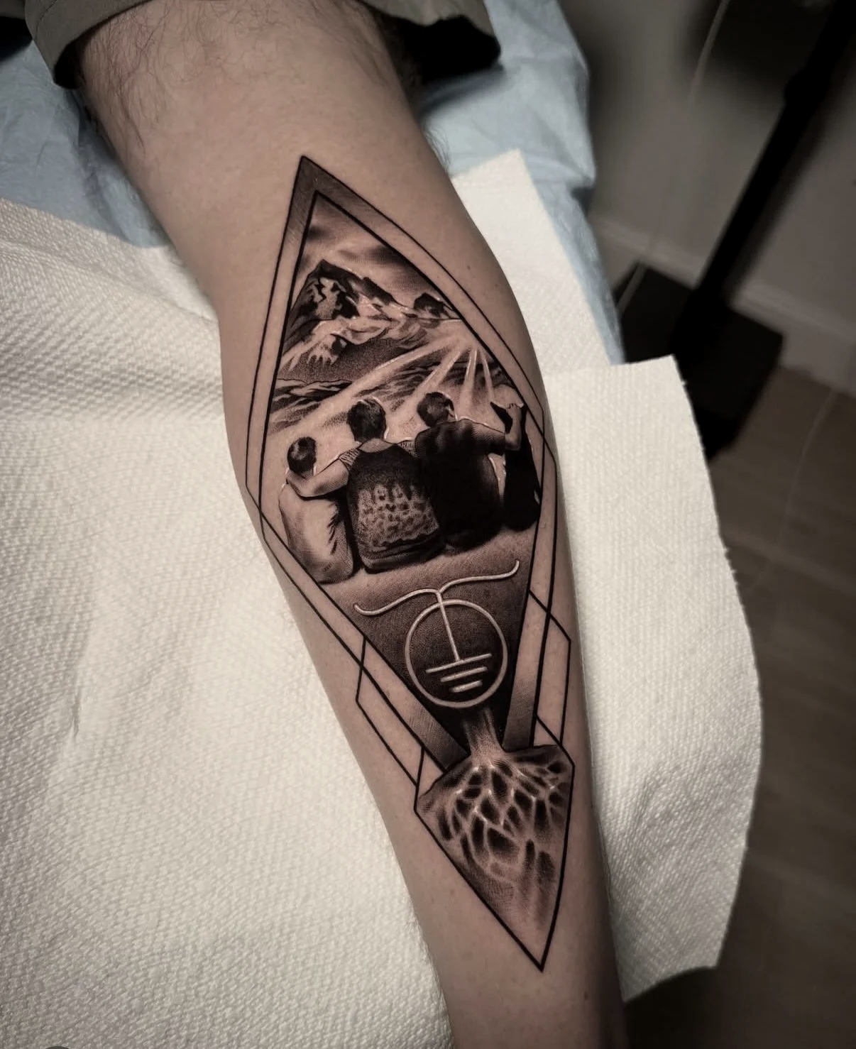 Tattoo of three children sitting together on a hill, watching a mountain landscape and sun rays.