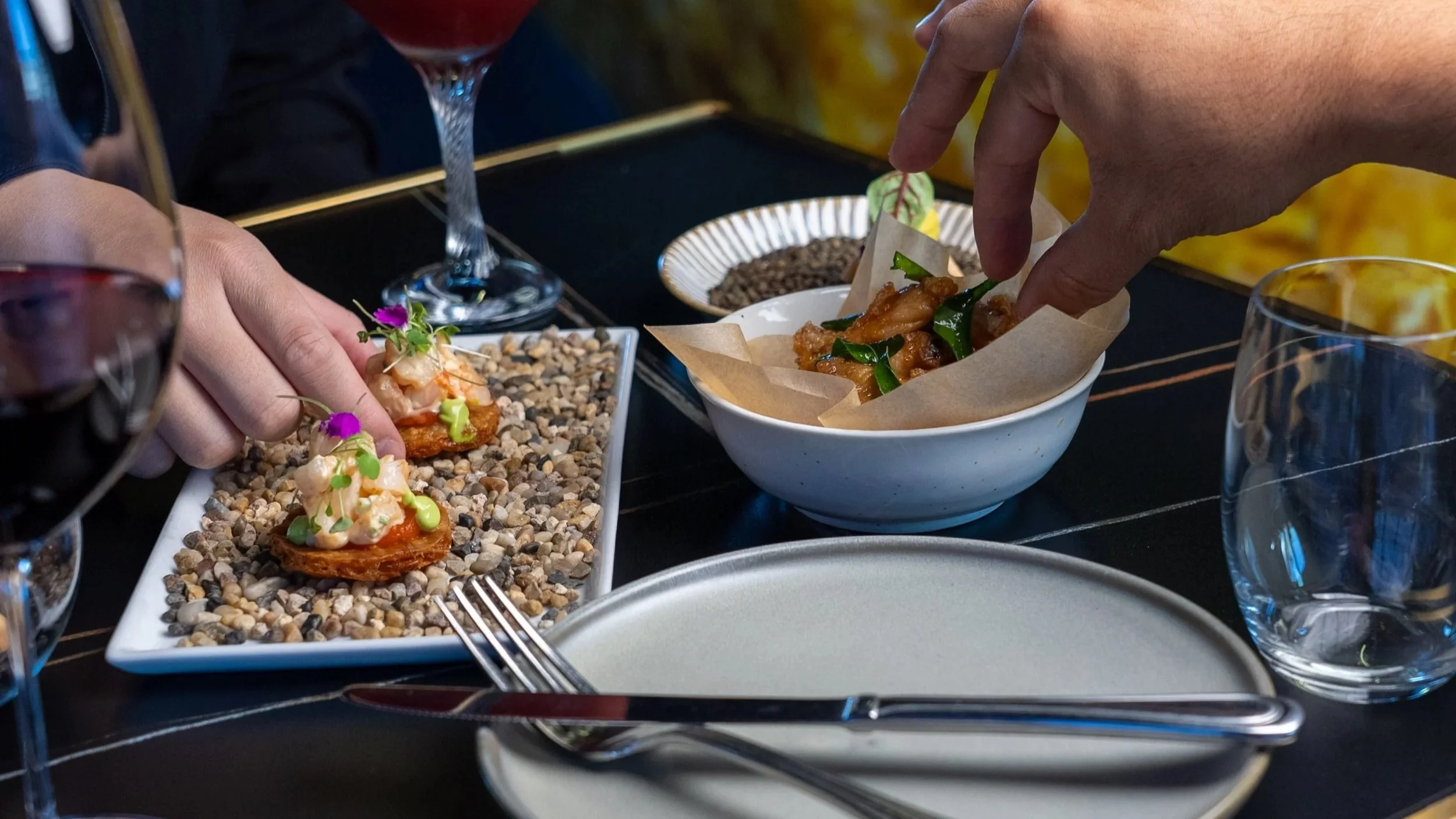 People enjoying a meal, with one person garnishing a dish with edible flowers on small appetizers arranged on a rectangular plate with pebbles, and another person serving food from a bowl containing stir-fried dish with vegetables on a dark table.