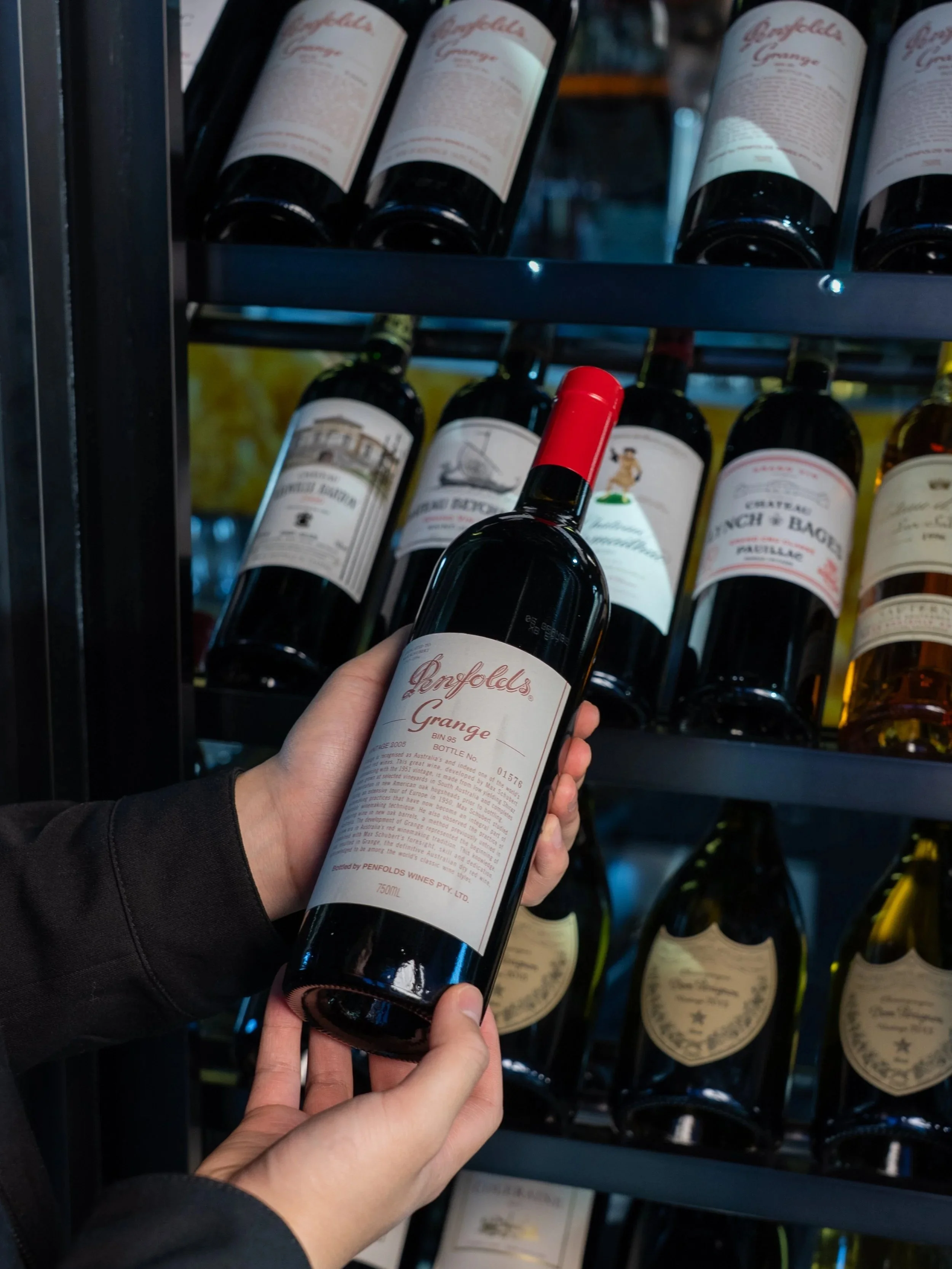 Person holding a bottle of Penfolds Grange red wine in front of a wine display shelf.