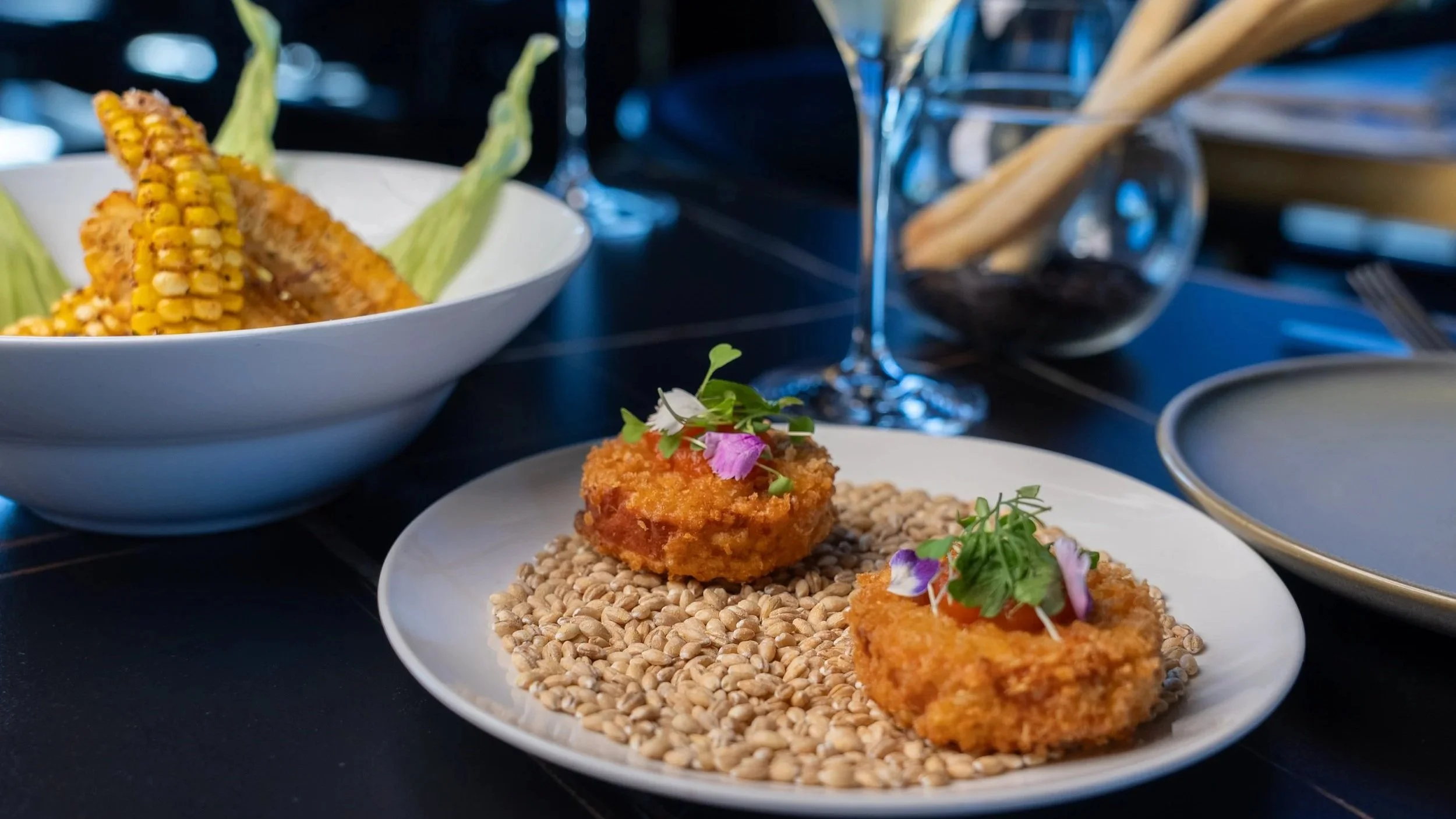 Two breaded and fried appetizers garnished with microgreens and edible flowers on a white serving plate, with a bed of toasted sesame seeds underneath, set on a black table surrounded by various dishes and drinks.