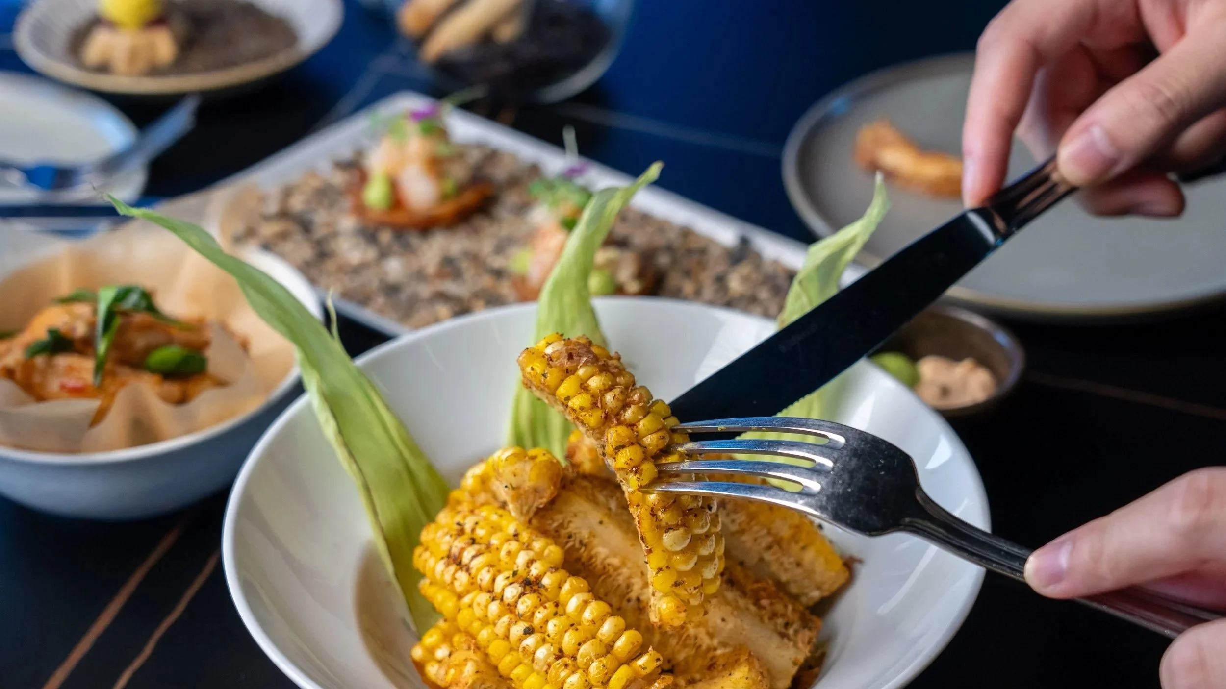 Person holding a fork and knife cutting into grilled corn on the cob in a white bowl, with other dishes of various foods on a dark table in the background.