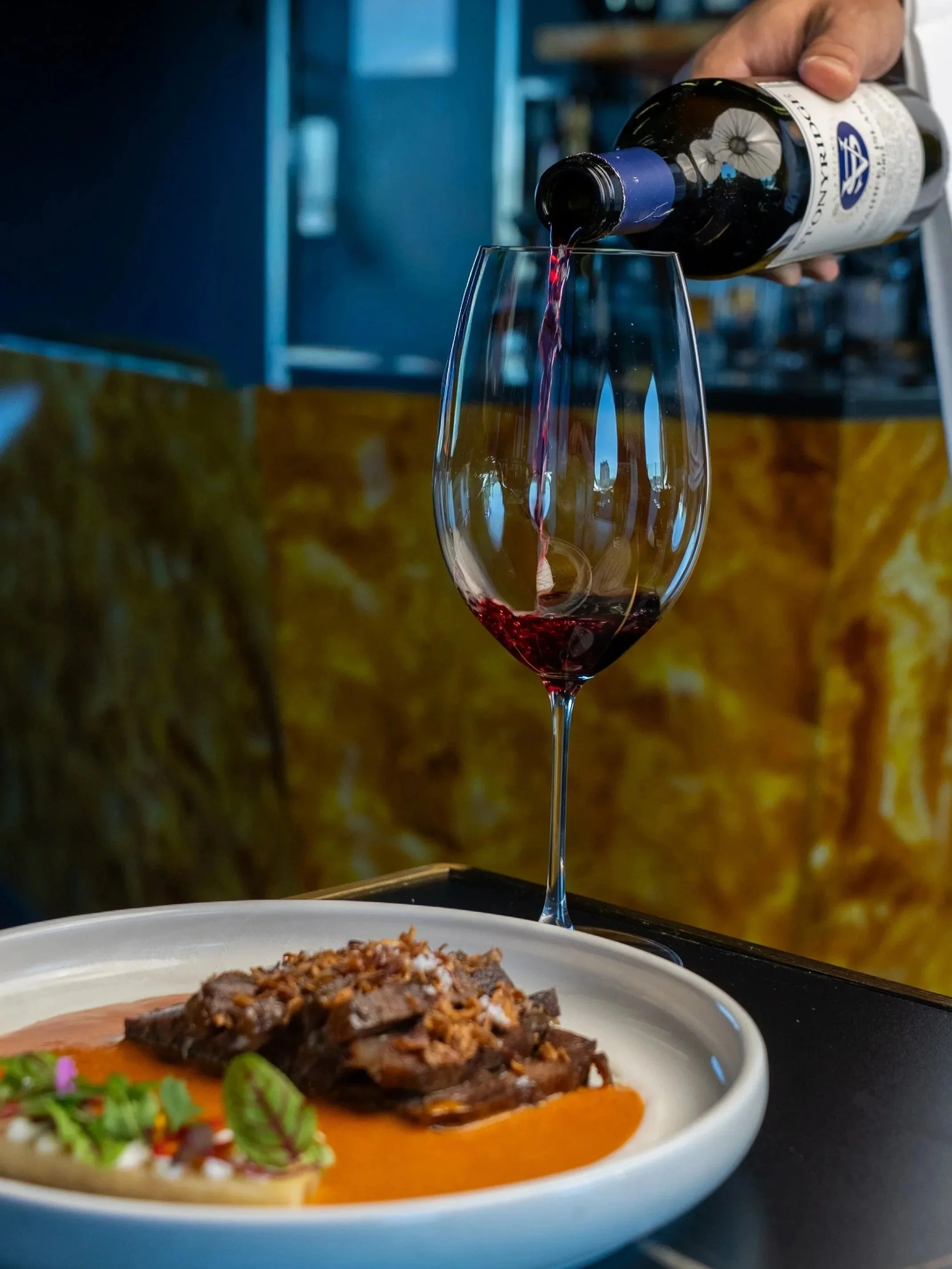 A hand pouring red wine into a wine glass above a plate of food on a table.