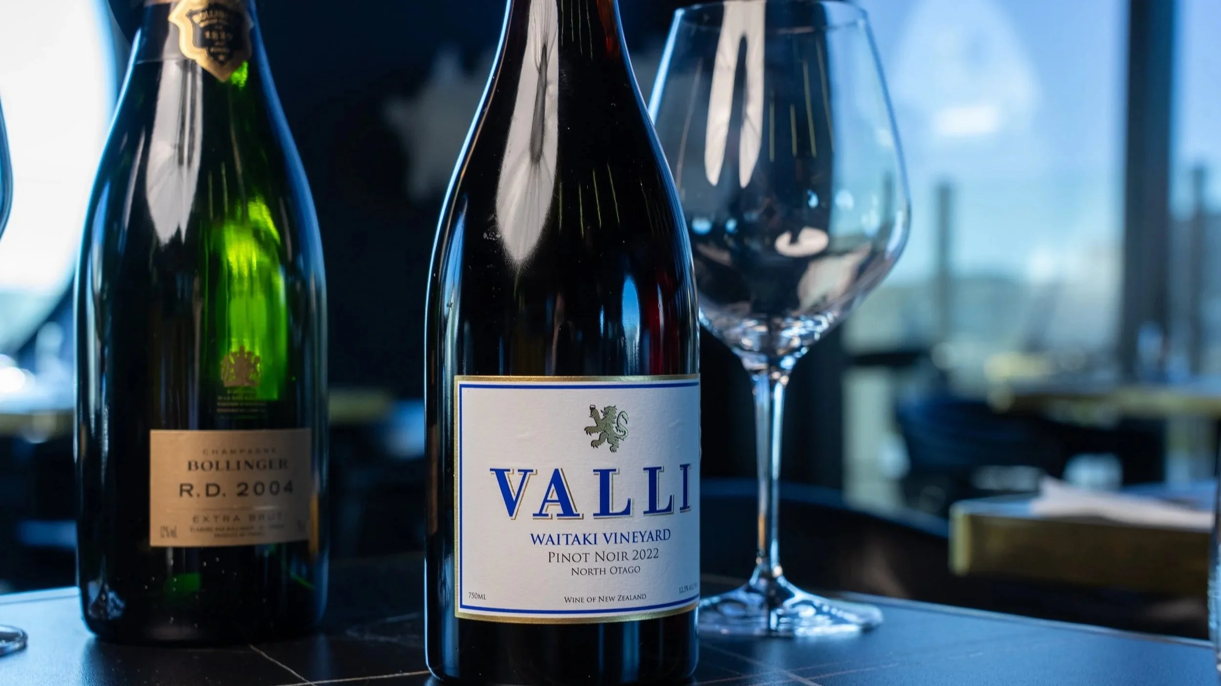 A bottle of Valli Waitaki Vineyard Pinot Noir wine from North Otago, New Zealand, accompanied by a glass of wine and another bottle of champagne in the background.