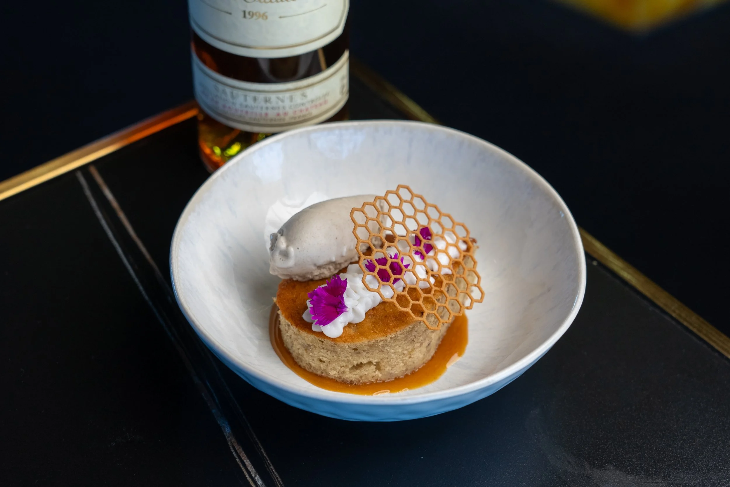 A dessert featuring a round cake topped with dollops of cream, edible purple flowers, a honeycomb-shaped caramel decoration, and a scoop of vanilla ice cream on a white bowl.