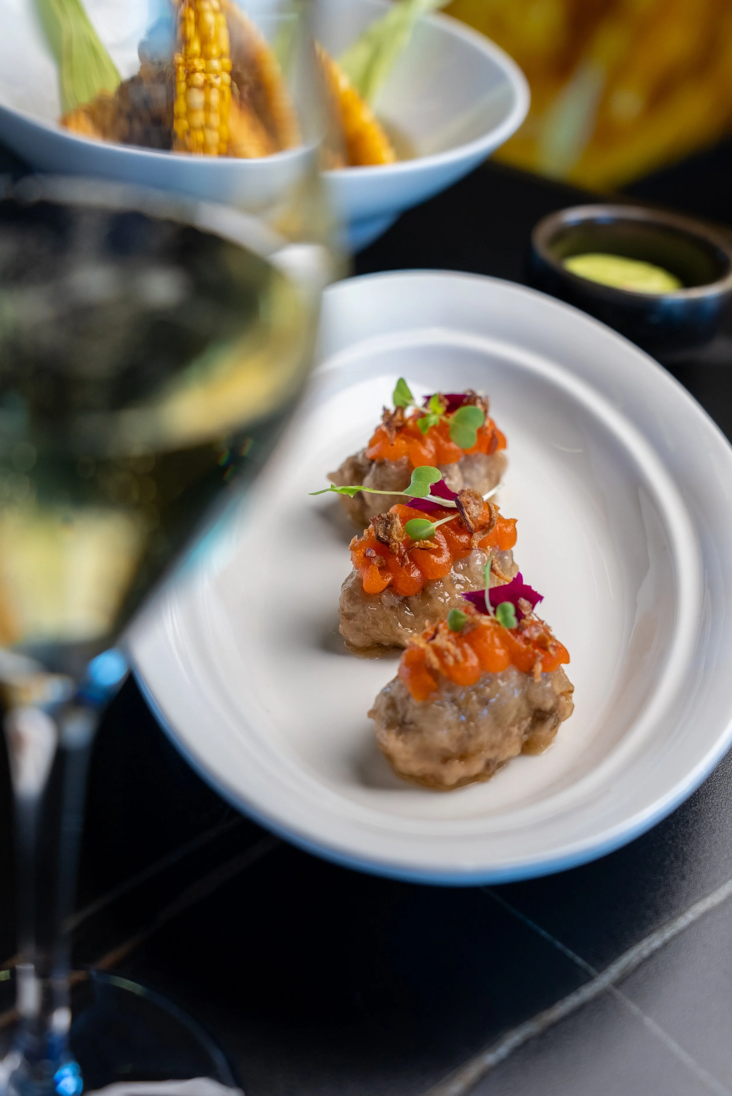 Three gourmet meatballs topped with orange bits and microgreens on a white oval plate, with a blurred glass of red wine and a bowl of corn in the background.