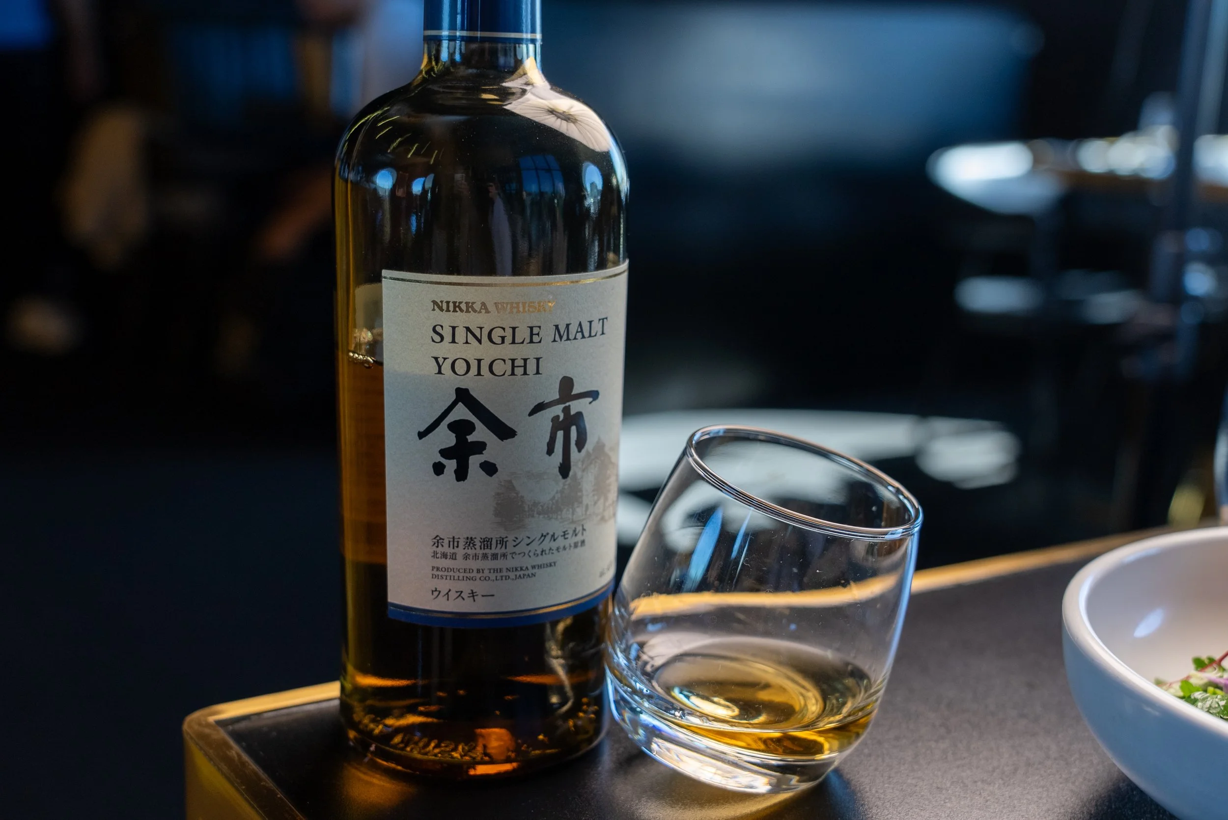A bottle of Nikka Whiskey Single Malt Yoichi with a glass of whiskey next to it, placed on a table, with a bowl partially visible on the right side.