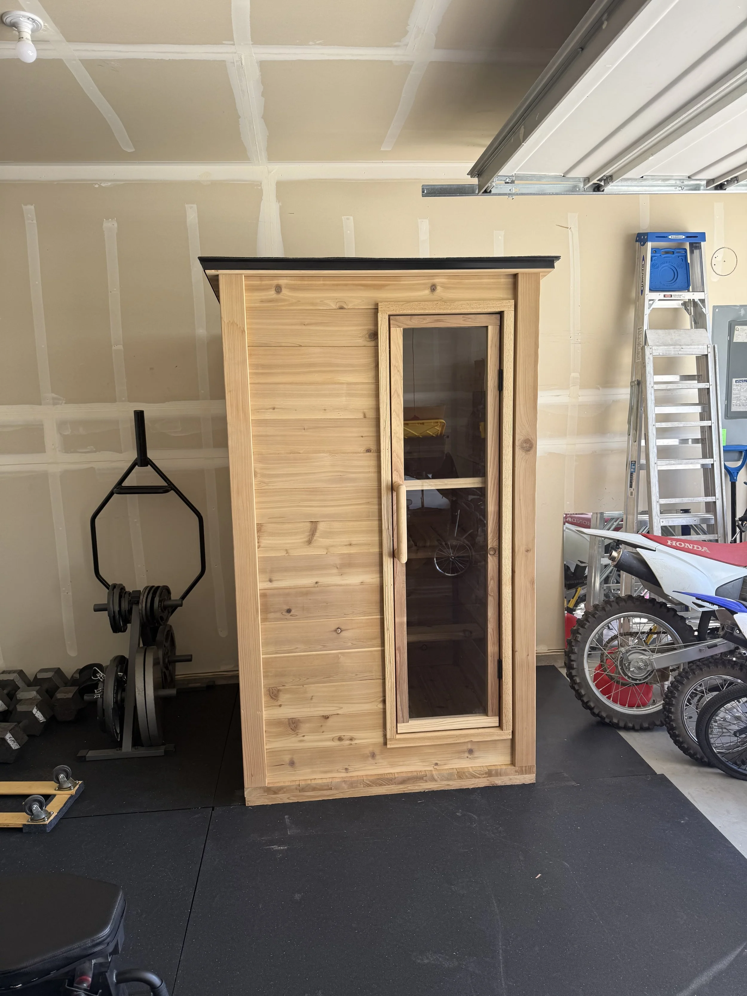 A garage with ceiling drywall installation, featuring a wooden cabinet with glass door, exercise equipment, a ladder, and a dirt bike.
