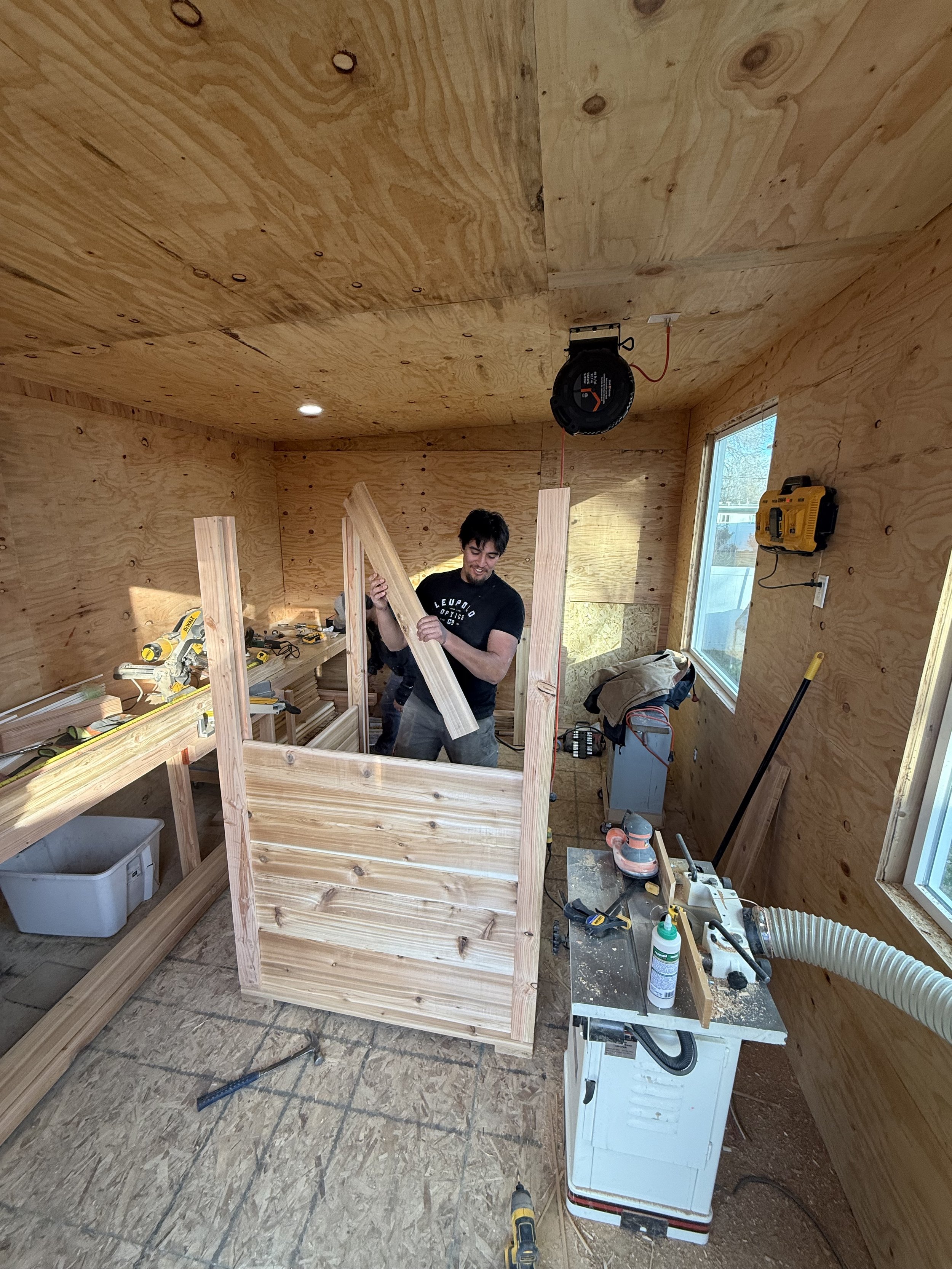 A man working on a woodworking project inside a small room with plywood walls and ceiling. Various tools and materials are visible, including a miter saw, a yellow tape measure, and wood pieces. The room has a window letting in natural light.