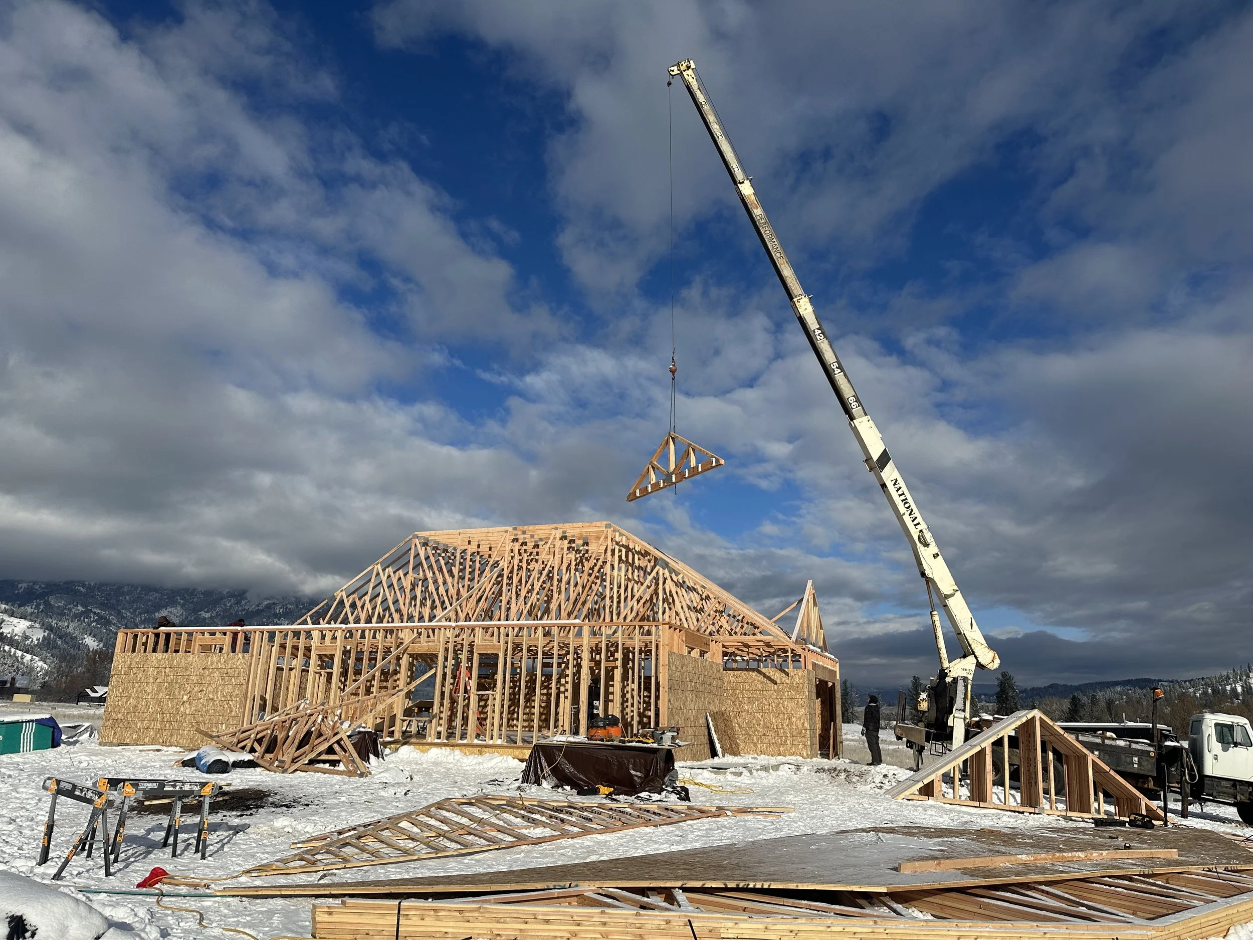 A construction site shows a wooden house frame under construction in a snowy landscape with mountains in the background. A crane is lifting a wooden roof truss into place, and construction workers are present, with building materials and tools scattered around.