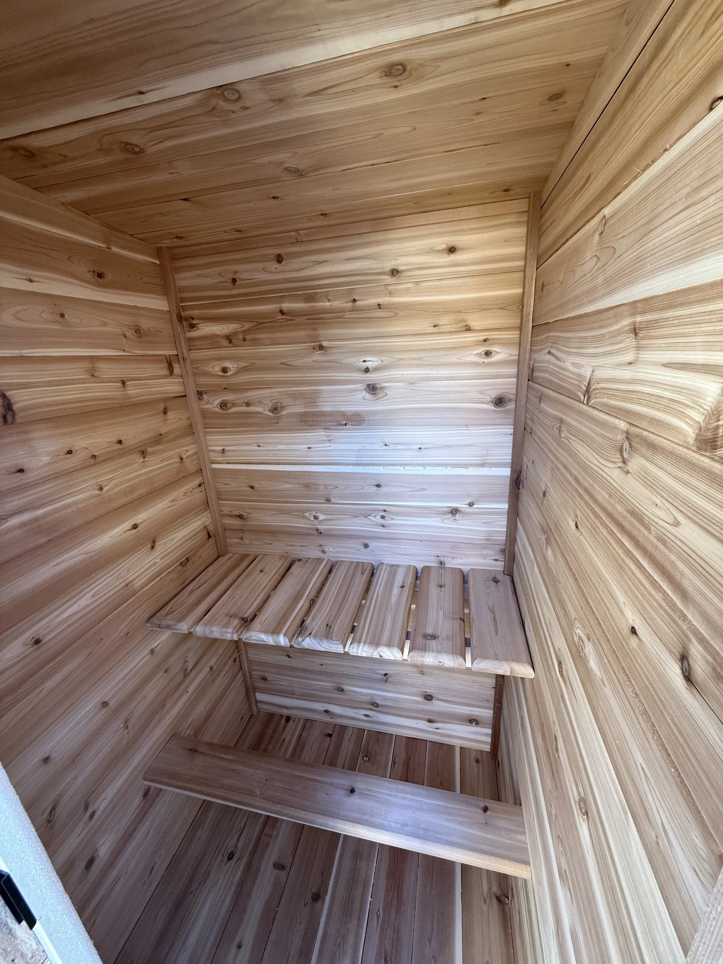 Interior view of a small wooden sauna with wooden walls, ceiling, and floor, featuring a wooden bench with slats for sitting.
