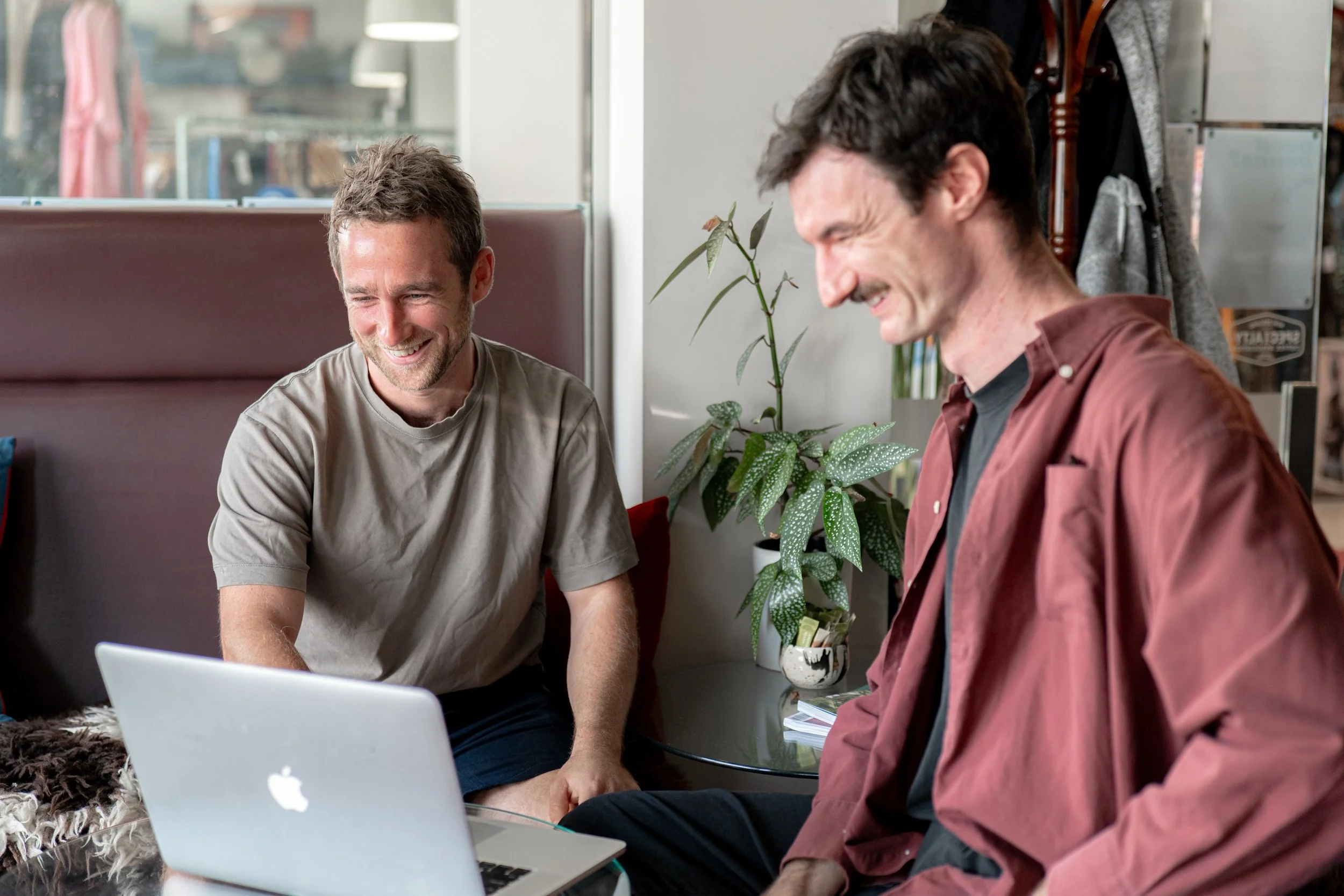 Two men sitting at a table, smiling and looking at a laptop, in a casual indoor setting with a plant nearby.
