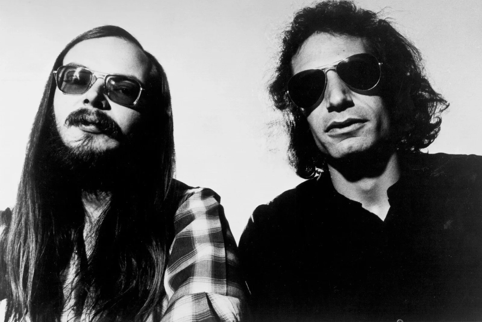 Black and white photo of two men wearing sunglasses, with long hair and beard, all with serious facial expressions.