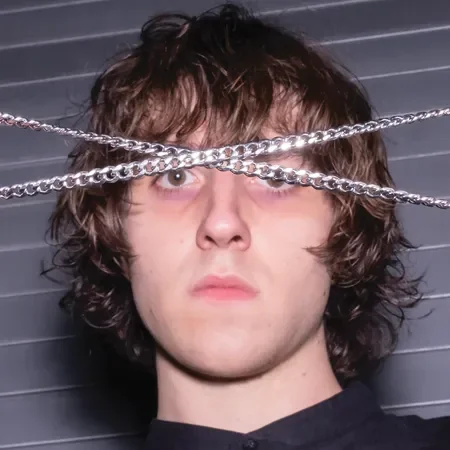Young man with messy brown hair, wide eyes, and a serious expression, with chains draped over his face in front of a dark, horizontal line background.