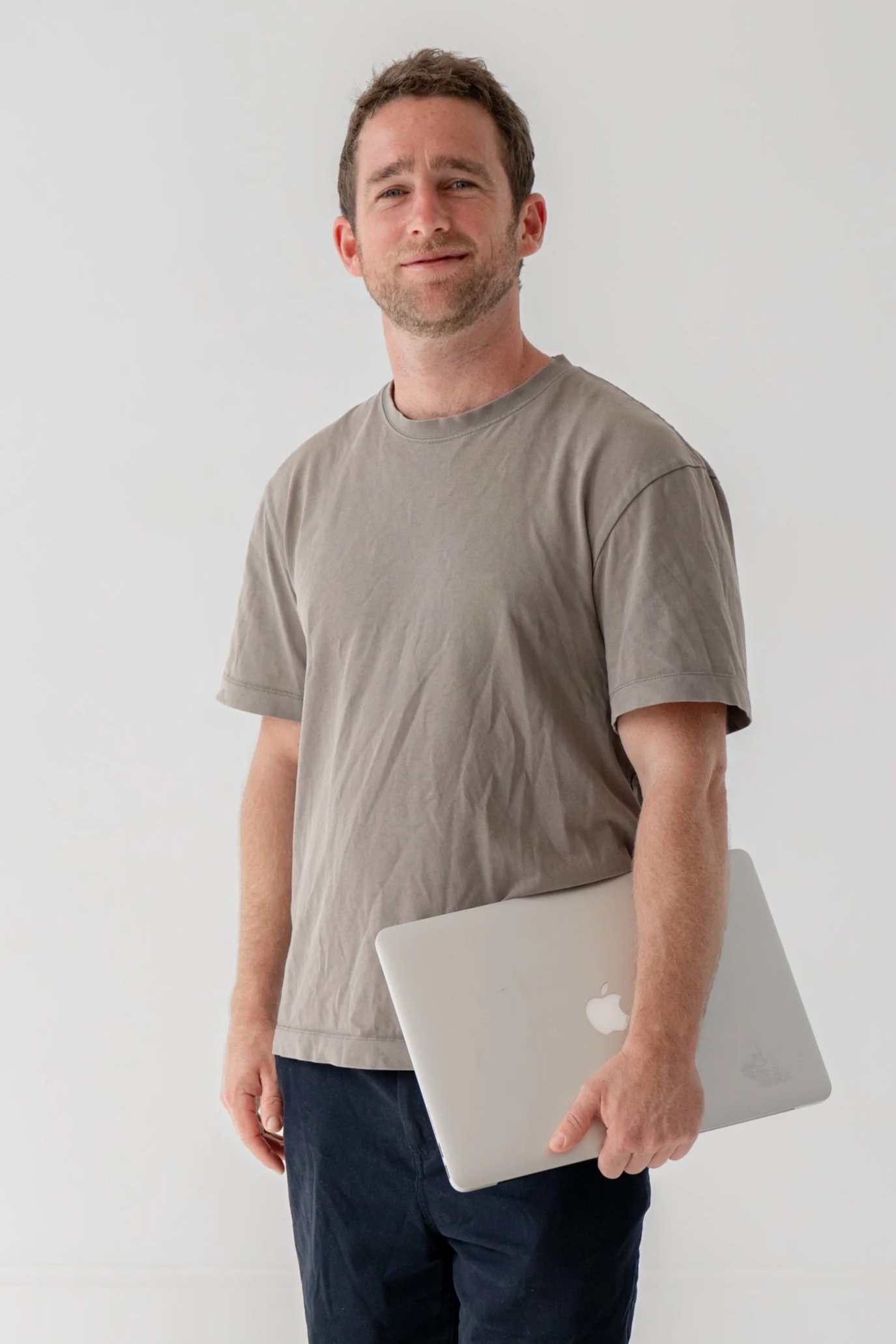 A man standing against a plain white wall, holding a silver laptop with an Apple logo, wearing a beige T-shirt and navy pants.