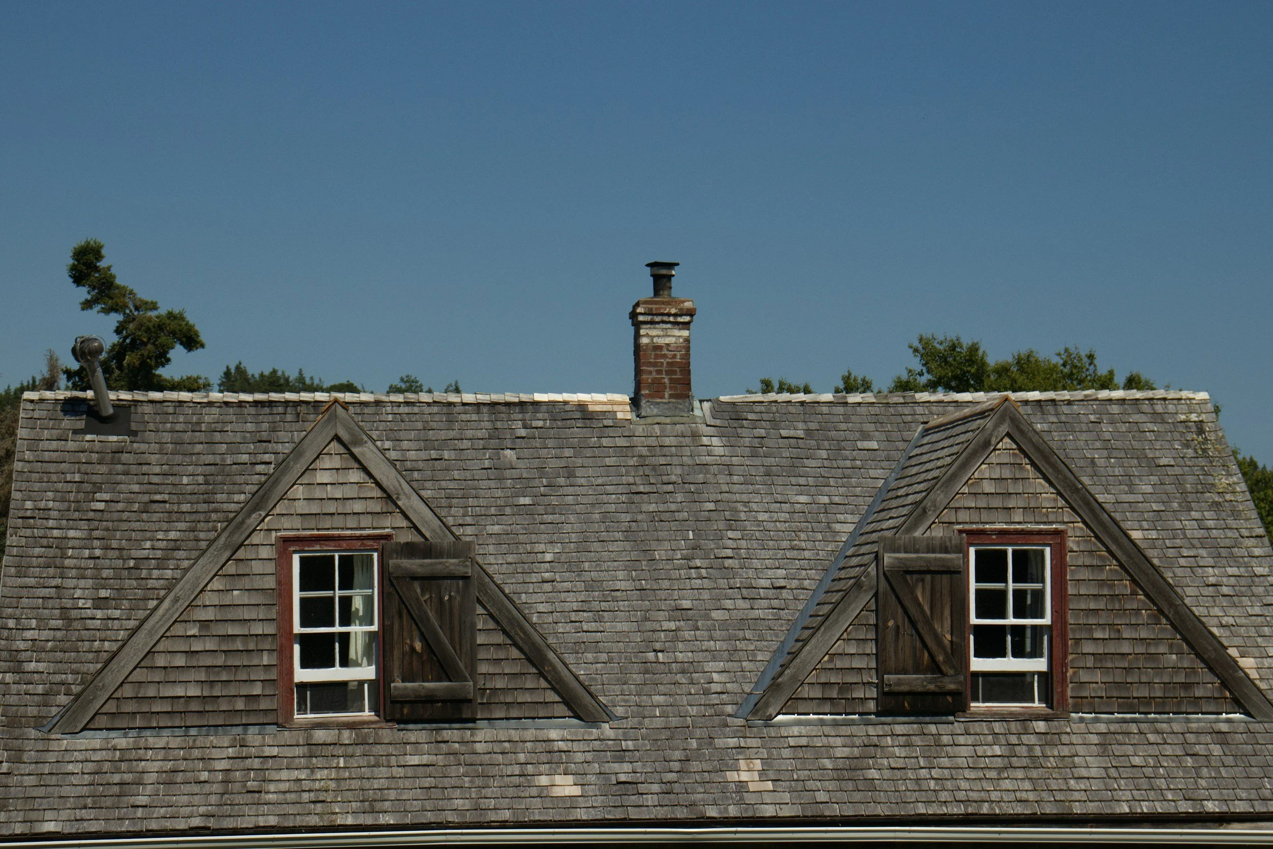 View of a house roof with two dormer windows, a chimney, and a vent pipe, under a blue sky with trees in the background.
