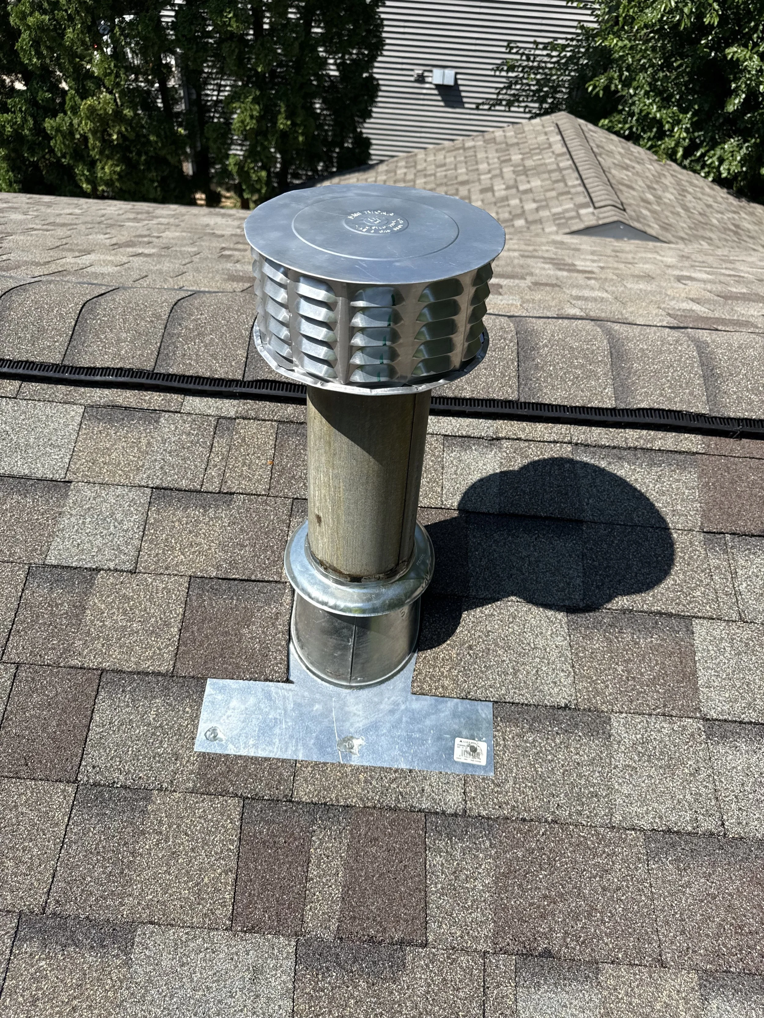 A roof vent pipe on a shingled roof with a metal cap, mounted on a wooden pipe with a metal flashing base visible, with trees and another roof in the background.