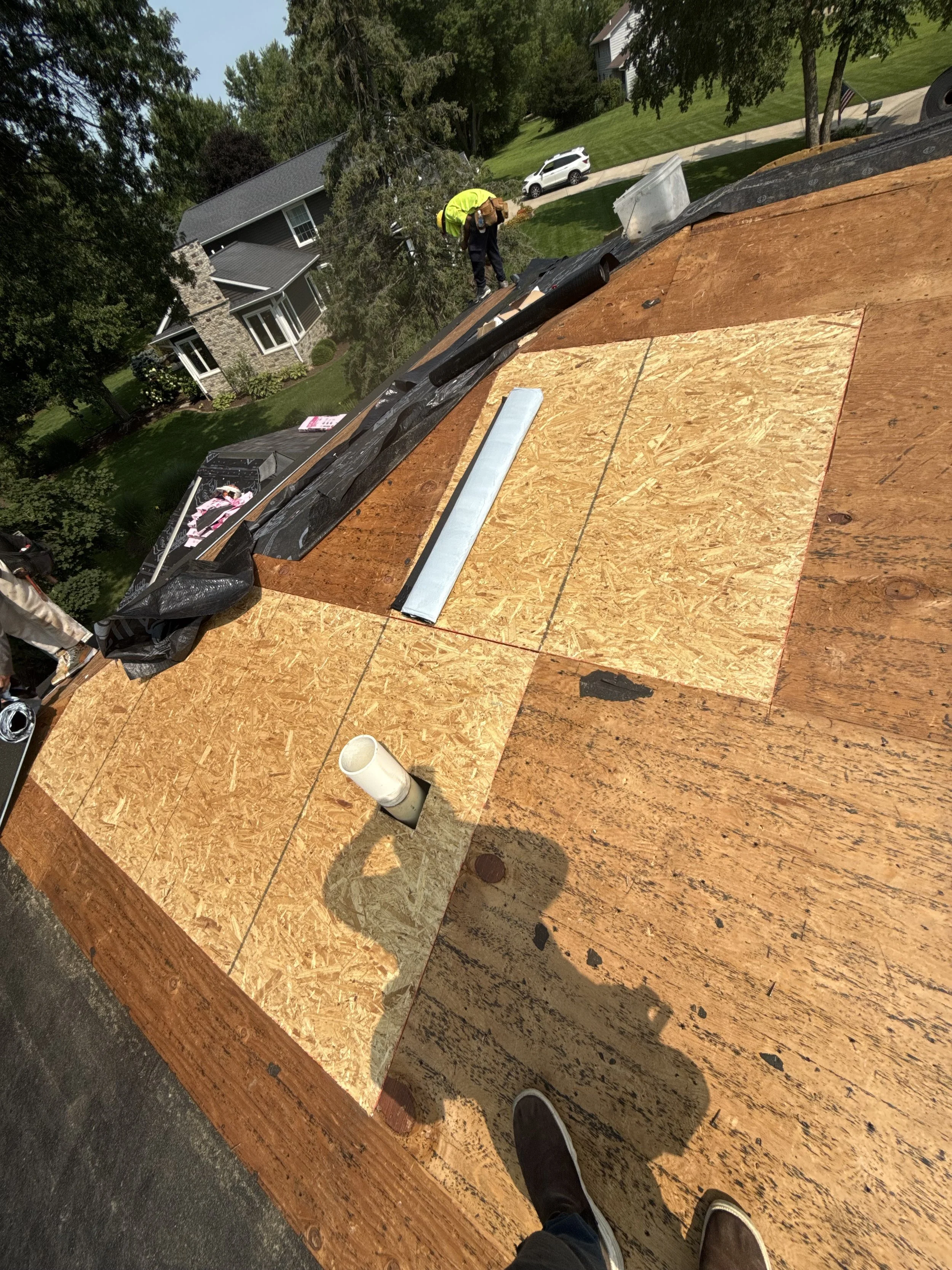 A worker on a roof under construction, with plywood sheets and roofing materials, surrounded by a residential neighborhood with trees and parked cars.