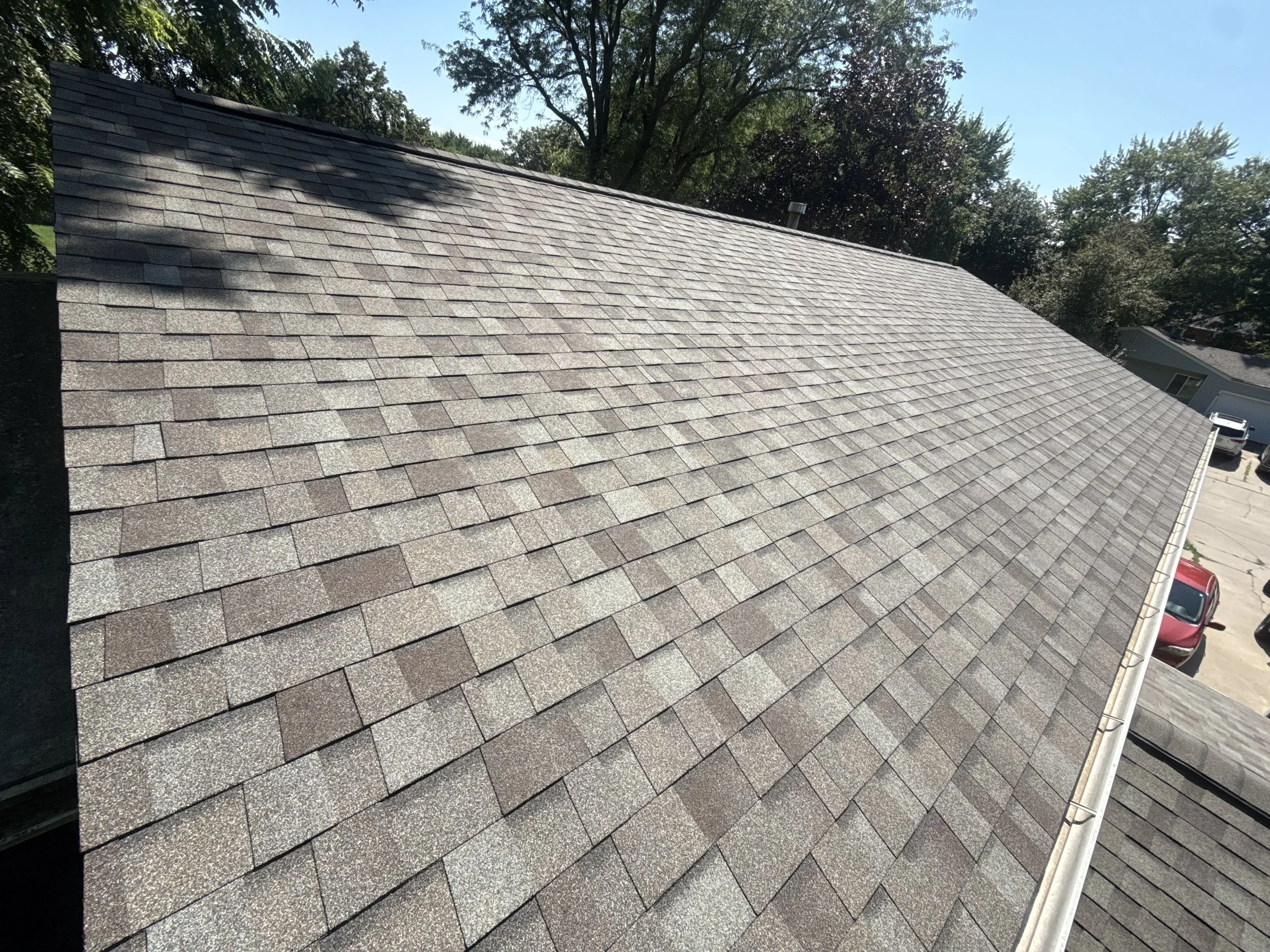 An aerial view of a sloped roof covered with gray asphalt shingles, with trees and parked cars visible in the background.