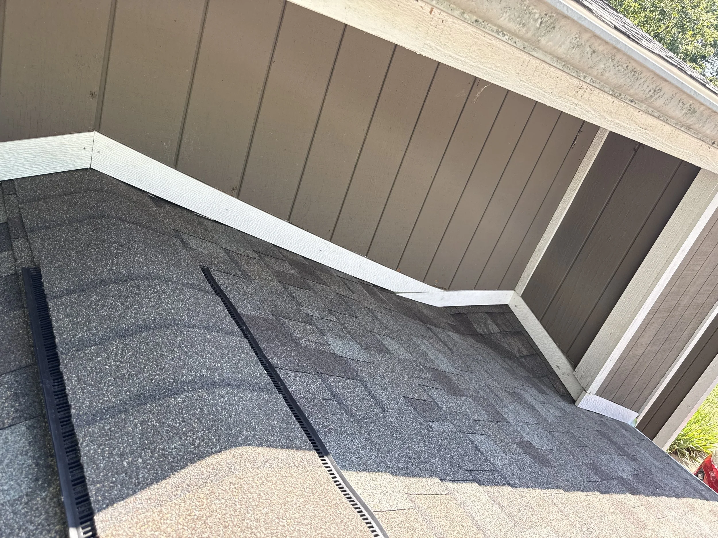Corner of a house roof with asphalt shingles, brown wooden siding, and white trim, with two black roof vents visible.