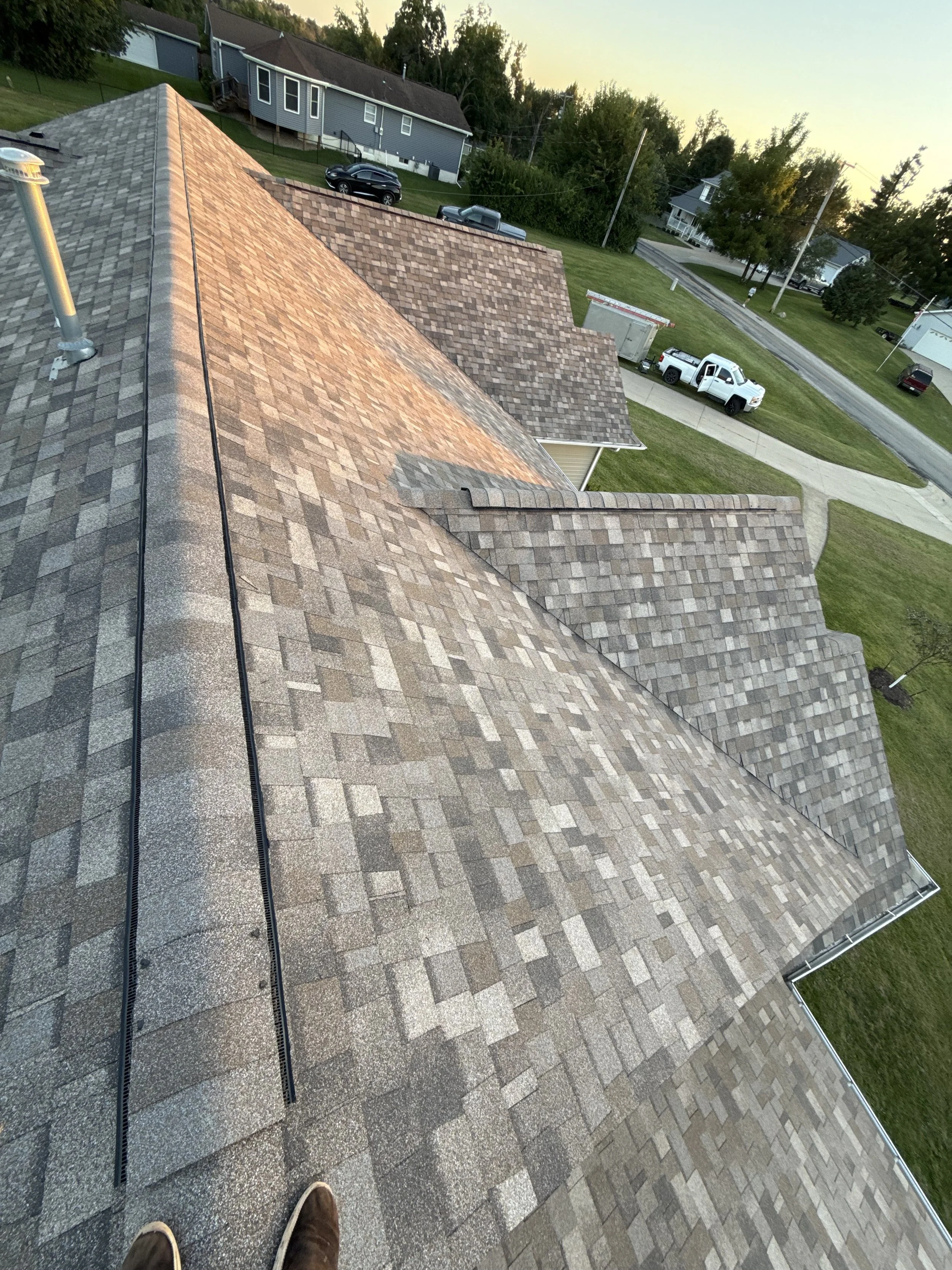 View of a residential roof with asphalt shingles, taken from a high vantage point at sunset. The roof has multiple sections and slopes, with a vent pipe visible on the left. The yard below features a lawn, driveway, and several parked vehicles, including a white truck with a utility bed.