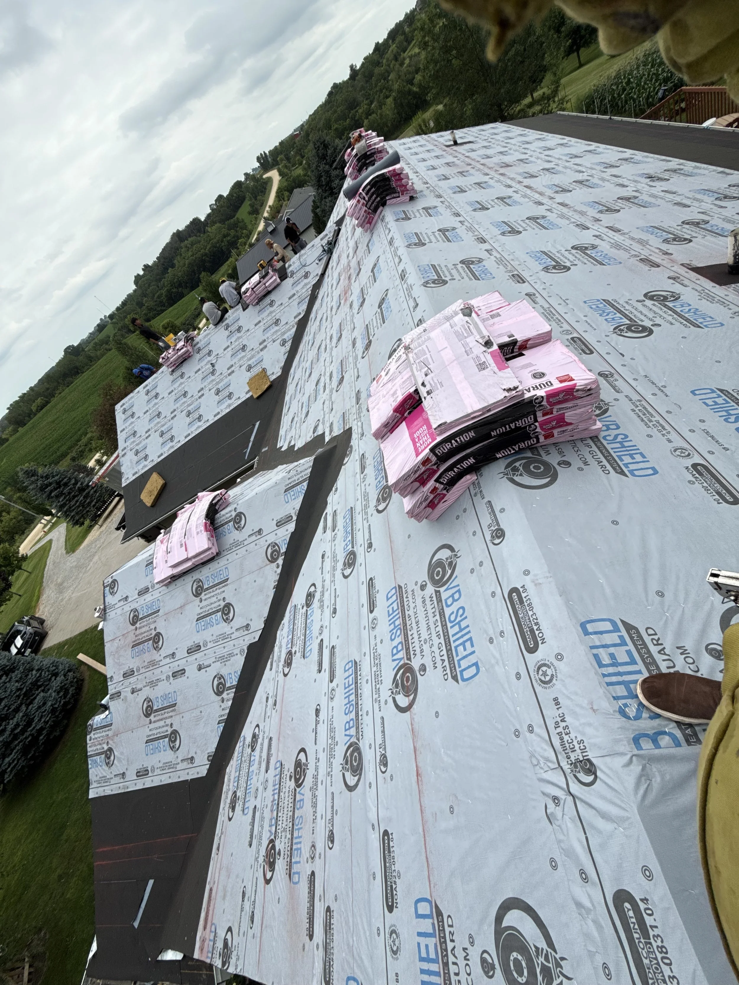 View of a building roof under construction with weatherproofing underlayment, shingles, and scaffolding, with workers and Lunds tools, against a background of green trees and cloudy sky.