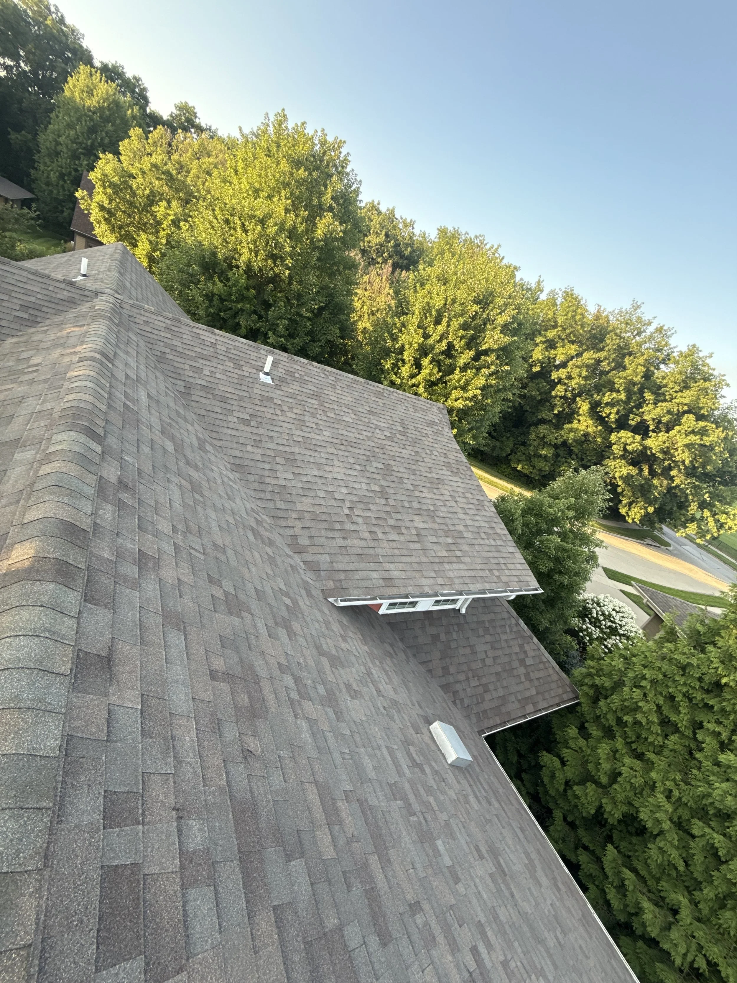 Aerial view of a house roof with brown shingles, surrounded by green trees and a neighborhood street.