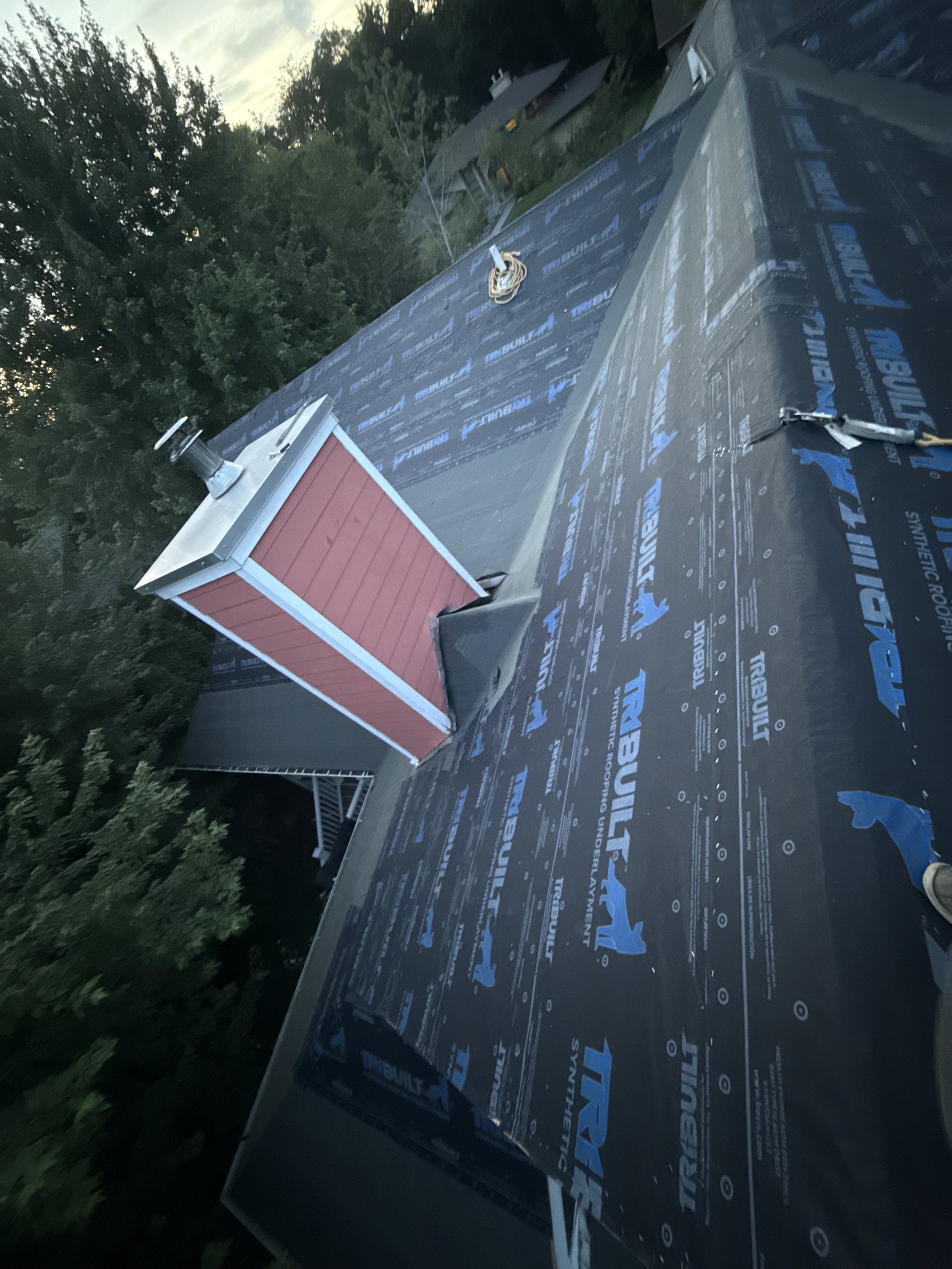 Roof under construction with black weatherproof underlayment labeled 'TrebuilT Synthetic Roofing Underlayment', a small chimney with red siding and white trim, and rooftop vents, with trees and houses in the background.