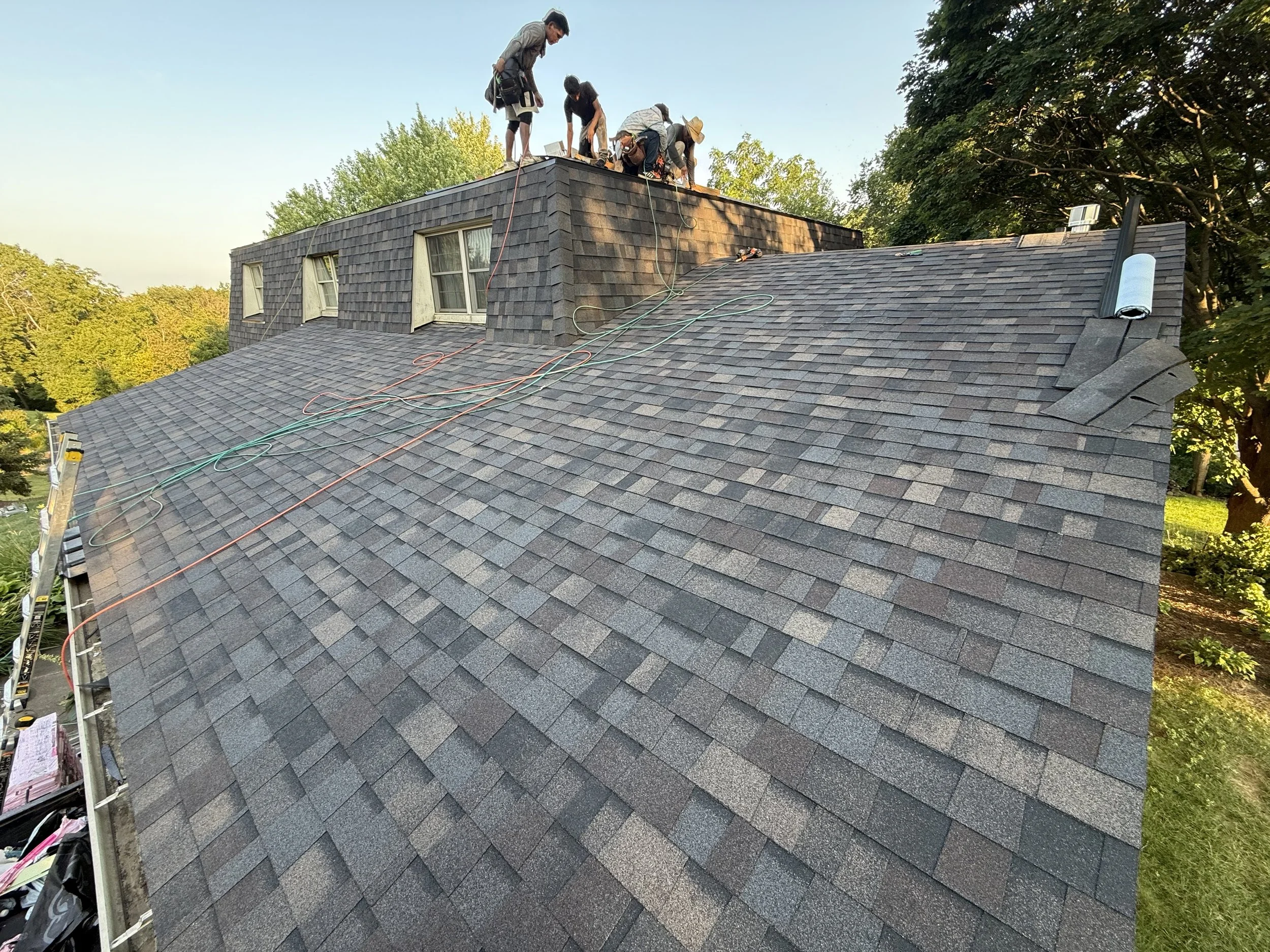 Roof repair workers on top of a house, working on the roof while standing near the edge, with tools and extension cords visible. The house has dark asphalt shingles, and the scene is surrounded by green trees.