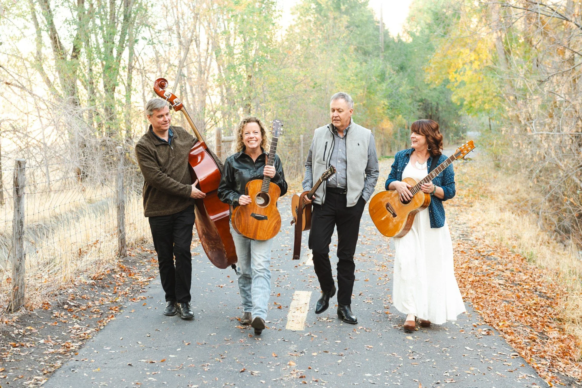 A group of five friends walking down a fall-colored forest road with fall leaves on the ground, smiling and holding musical instruments.