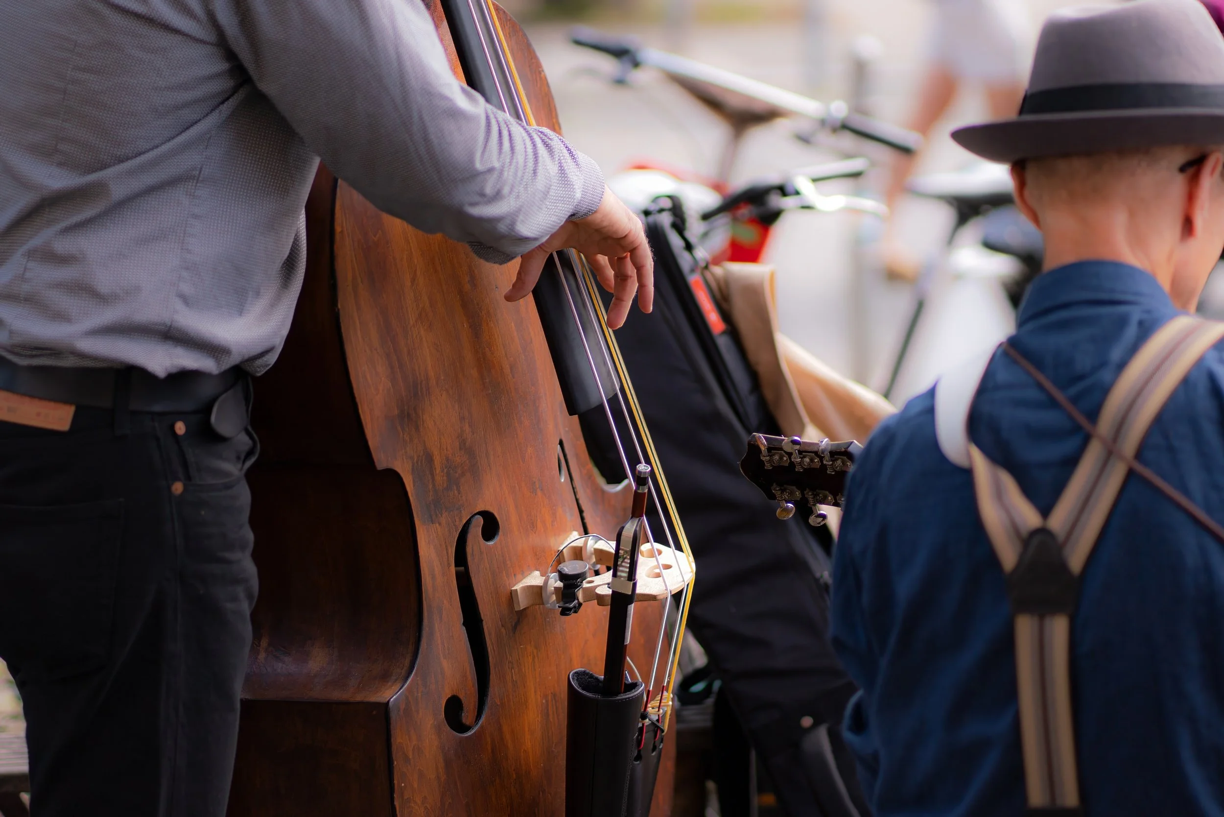 A person playing a double bass, with another person nearby wearing a hat, at an outdoor event.