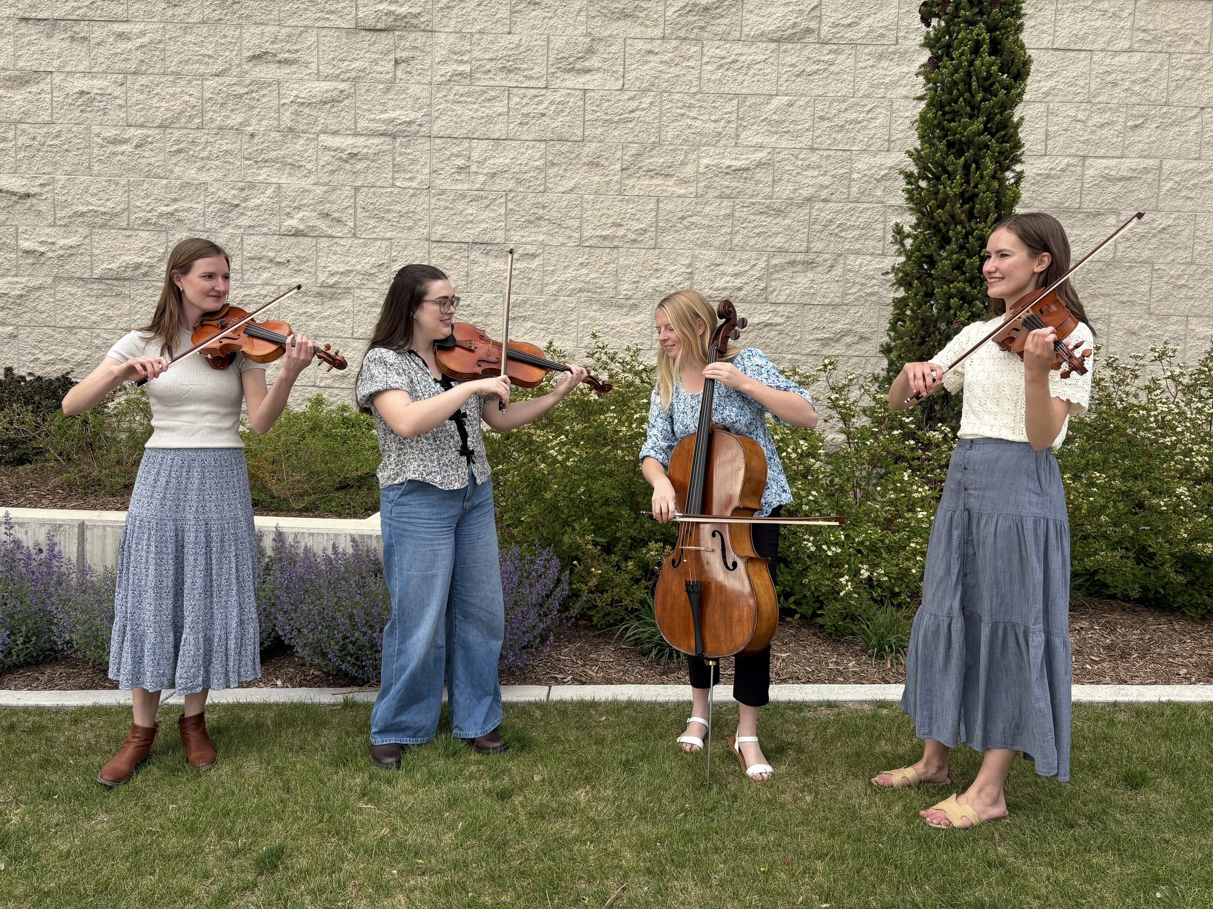 Four women playing string instruments outdoors in front of a stone wall and greenery, with one woman playing the cello and the others playing violins.
