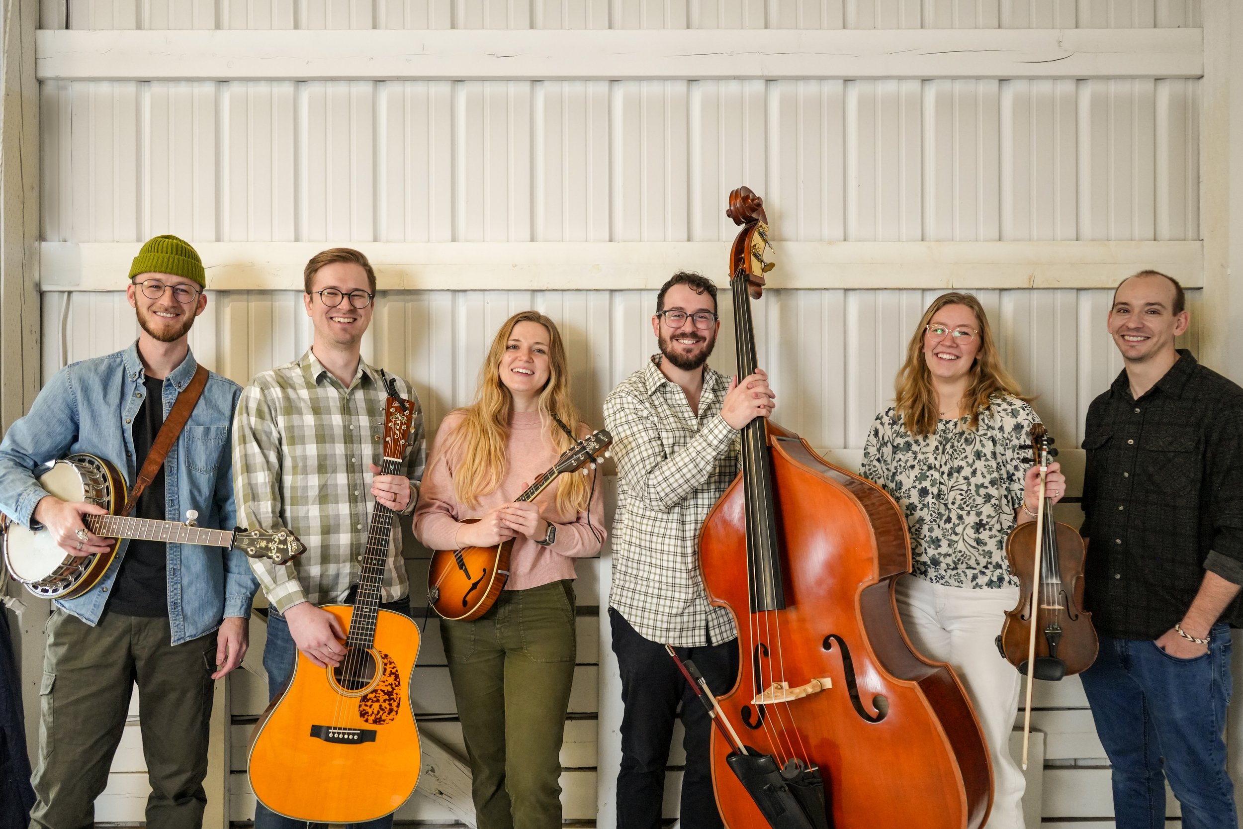 A diverse group of seven musicians holding various musical instruments, smiling and standing against a wood-paneled wall.