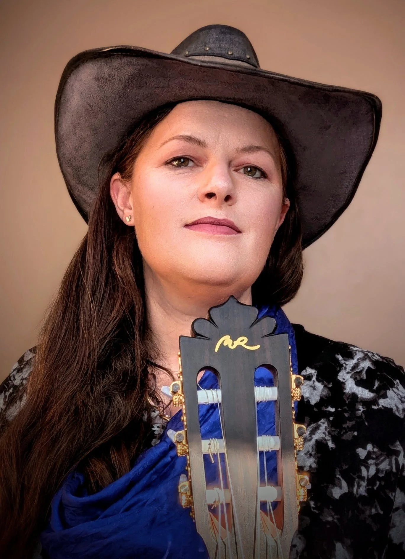 Woman wearing a black cowboy hat with metal embellishments, black and white patterned jacket, and blue dress, holding a decorative cutout with the word 'Mrs.' at a celebration or event.