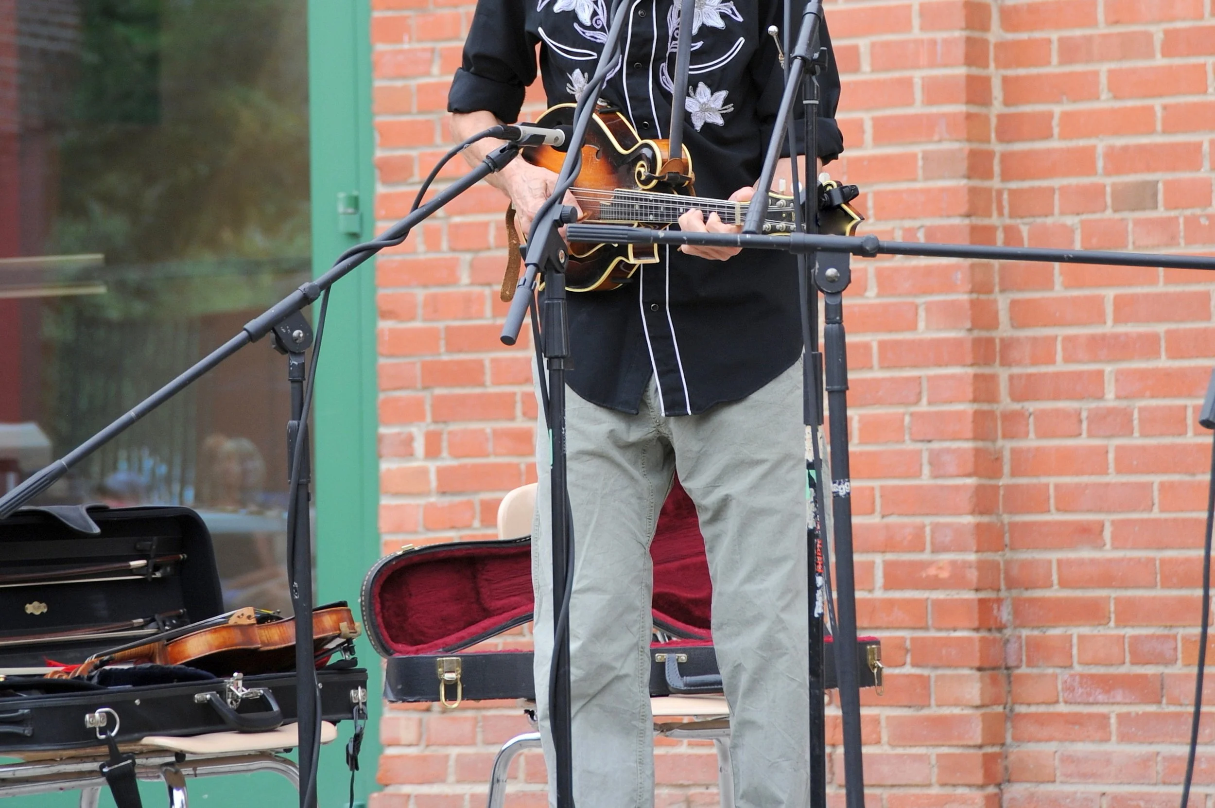 A person playing a small stringed instrument, possibly a mandolin or violin, outdoors in front of a brick wall. The person’s face is not visible, and they are dressed in a black shirt with floral embroidery and light-colored pants. Various musical equipment is around them, including a violin case and microphone stand.